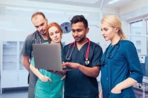 Four medical professionals in scrubs review a laptop. Inside a medical room, two men and two women look closely.