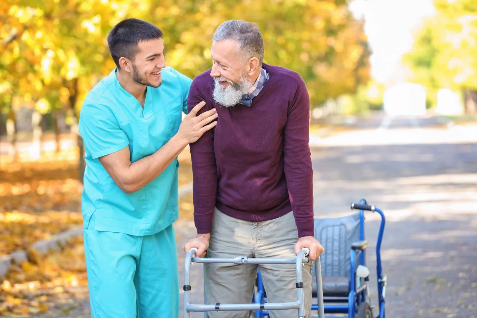 Man in turquoise scrubs helps an older man walk with a walker, both smiling, outdoors.