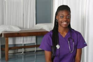 Smiling Black woman in purple scrubs with stethoscope in a medical setting.