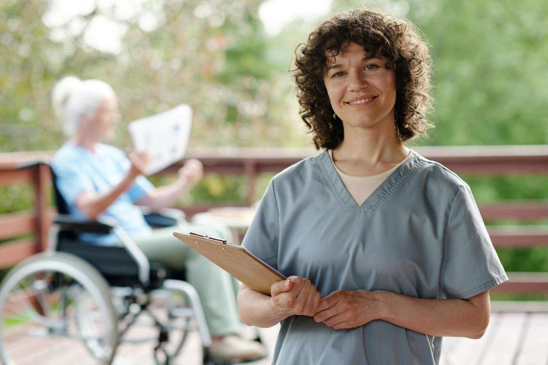 Caregiver smiling with clipboard beside a wheelchair user reading outdoors on a sunny patio