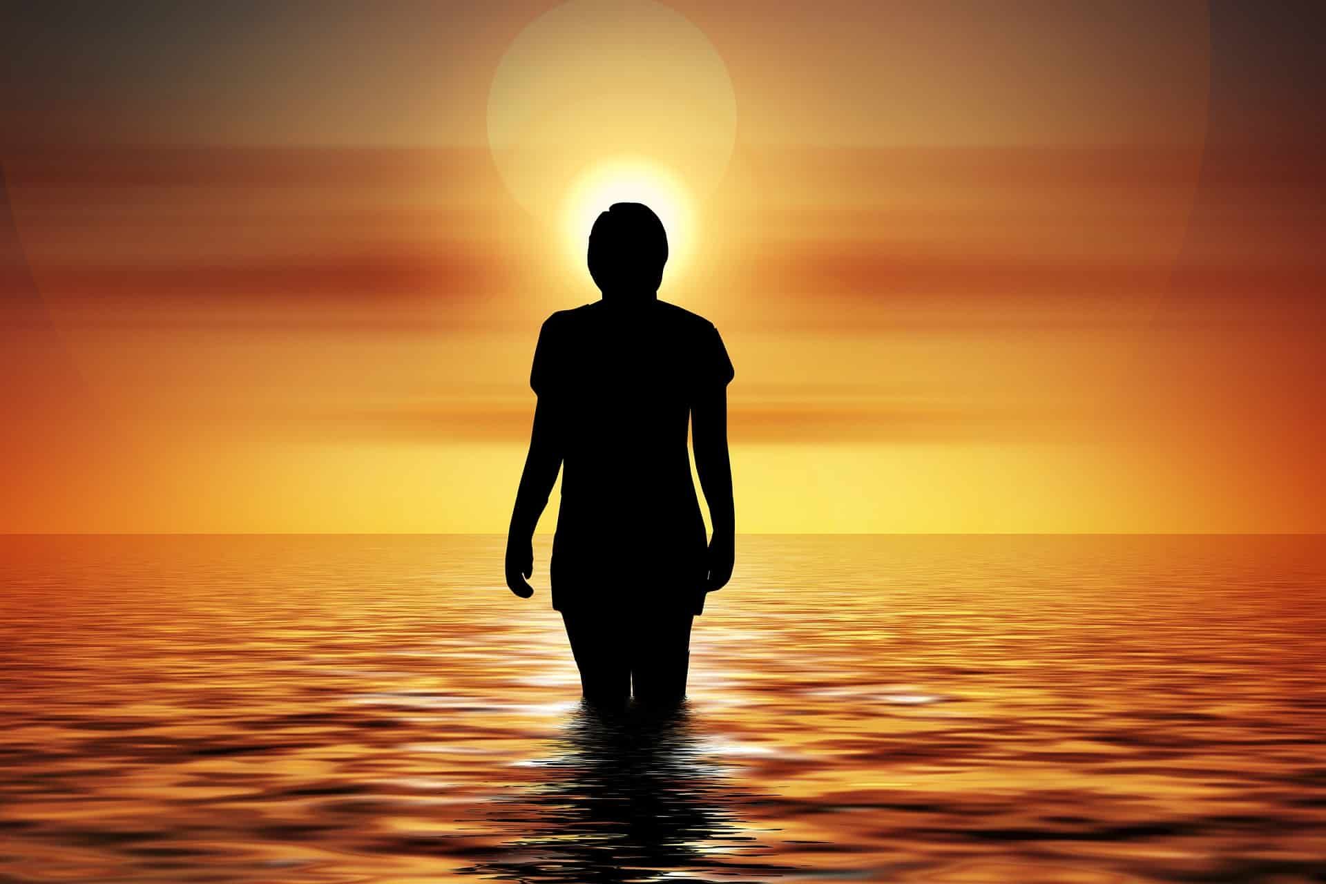 Silhouette of a person wading in water toward the setting sun, creating an orange and yellow gradient.