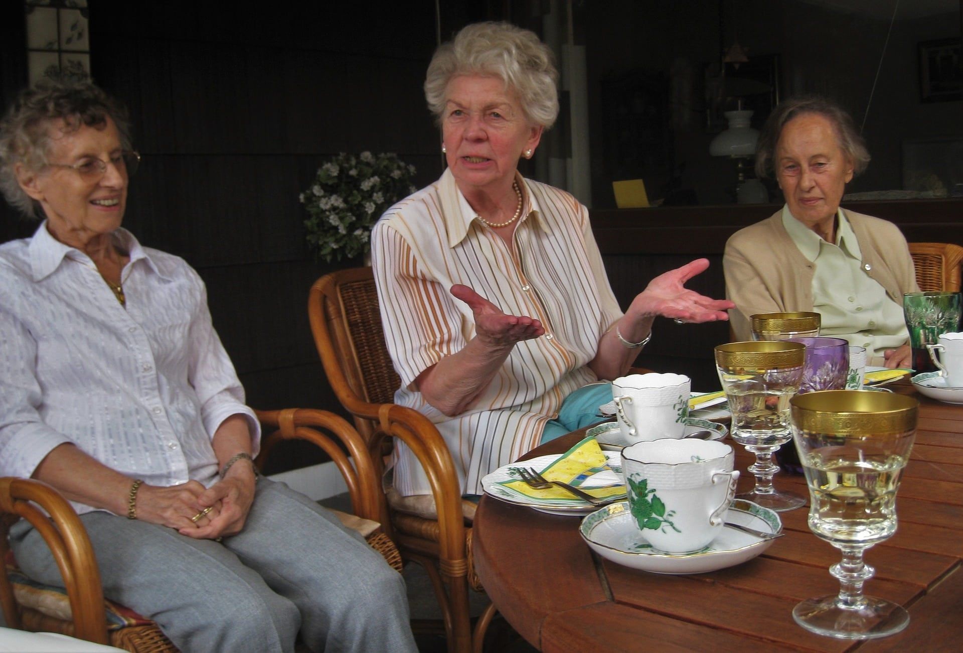 Three older women seated at a table, conversing; one gestures with hands. Table with drinks, outside setting.