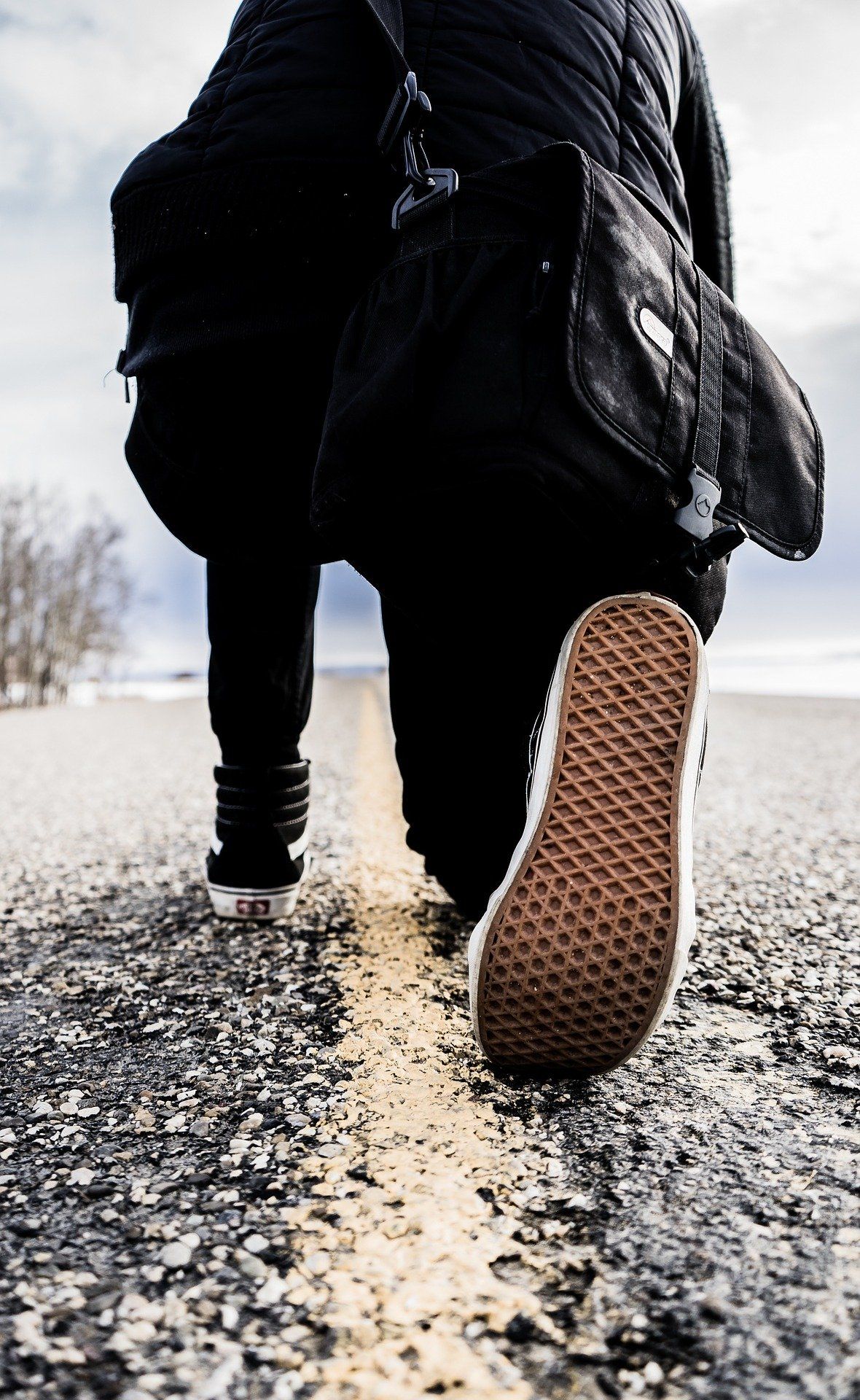 Person walking down a road, low-angle shot. Wearing black jacket, pants, and bag. Brown shoe sole visible.