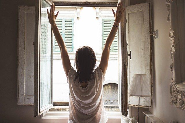Woman with arms raised in front of open window, reaching towards sunlight.