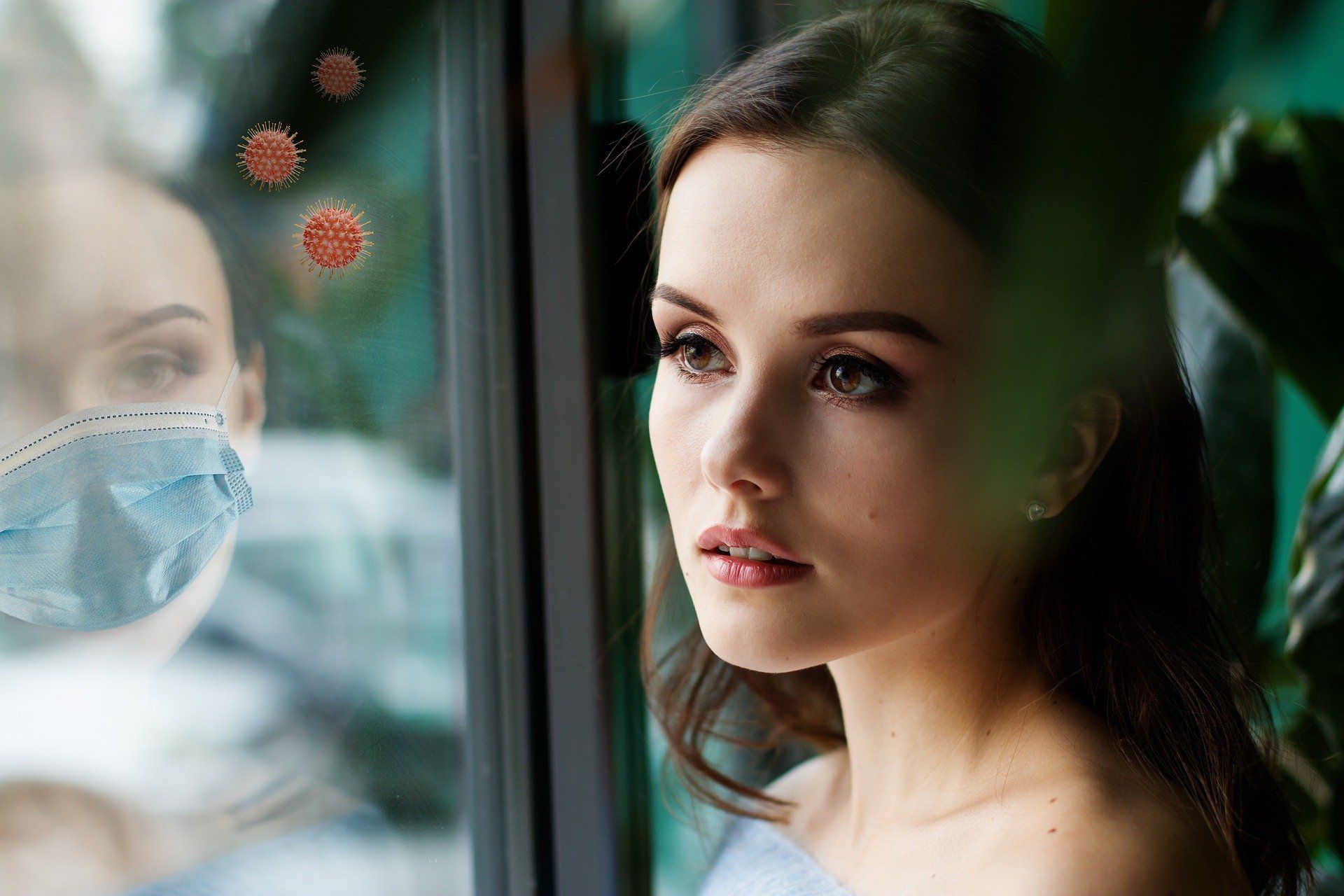 Woman looks out a window, seeing a reflection of a face mask and floating virus particles.