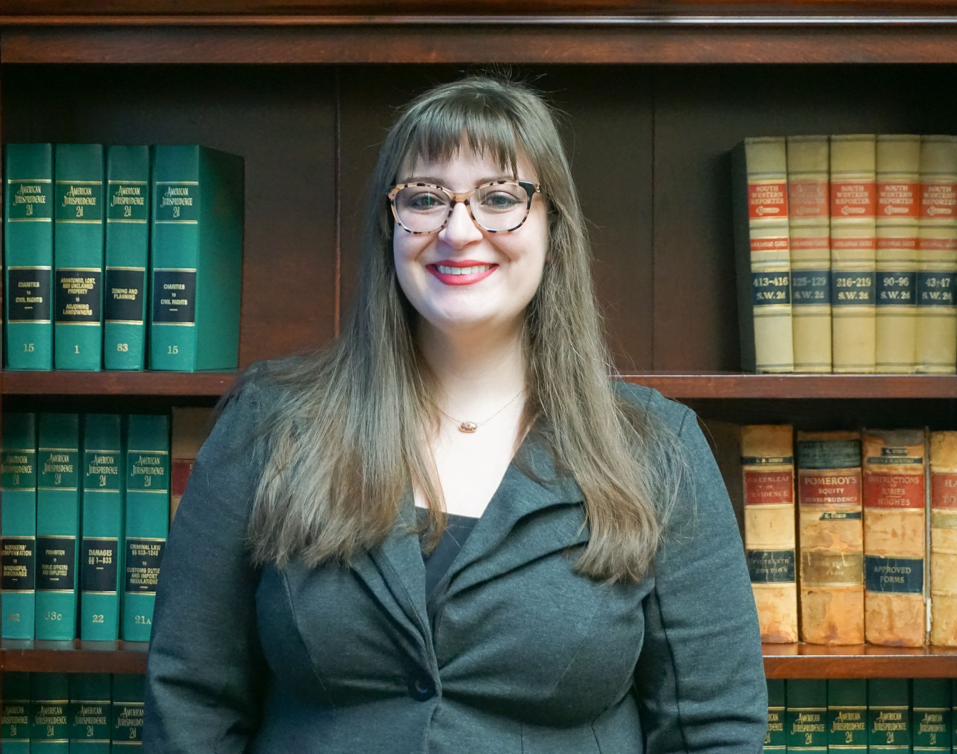 Woman in glasses and blazer smiles in front of a bookshelf filled with books.