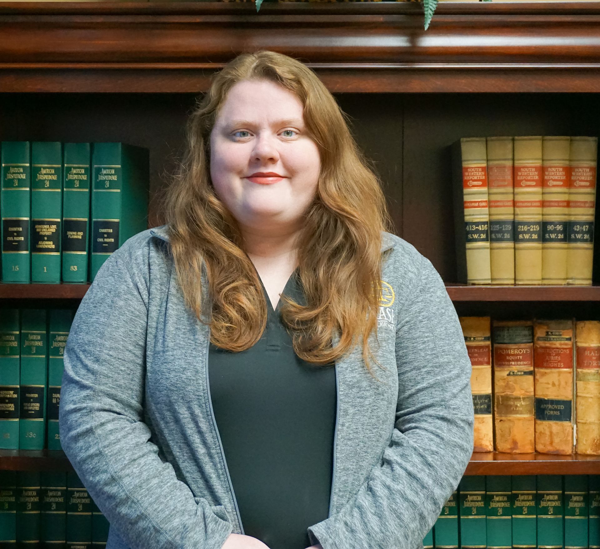 Woman with red hair, wearing a gray cardigan, standing in front of a bookshelf with law books.