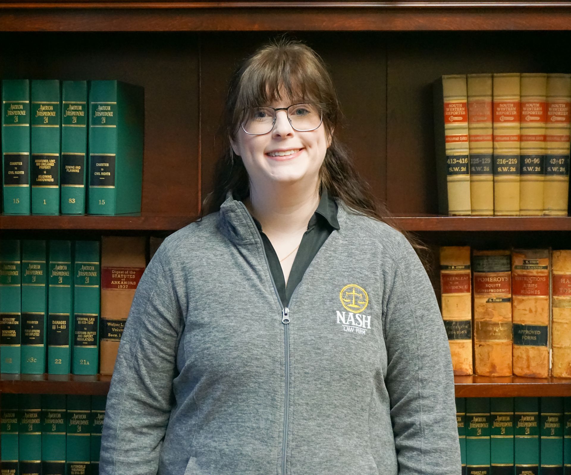 Woman in glasses and gray jacket smiles in front of a bookcase.