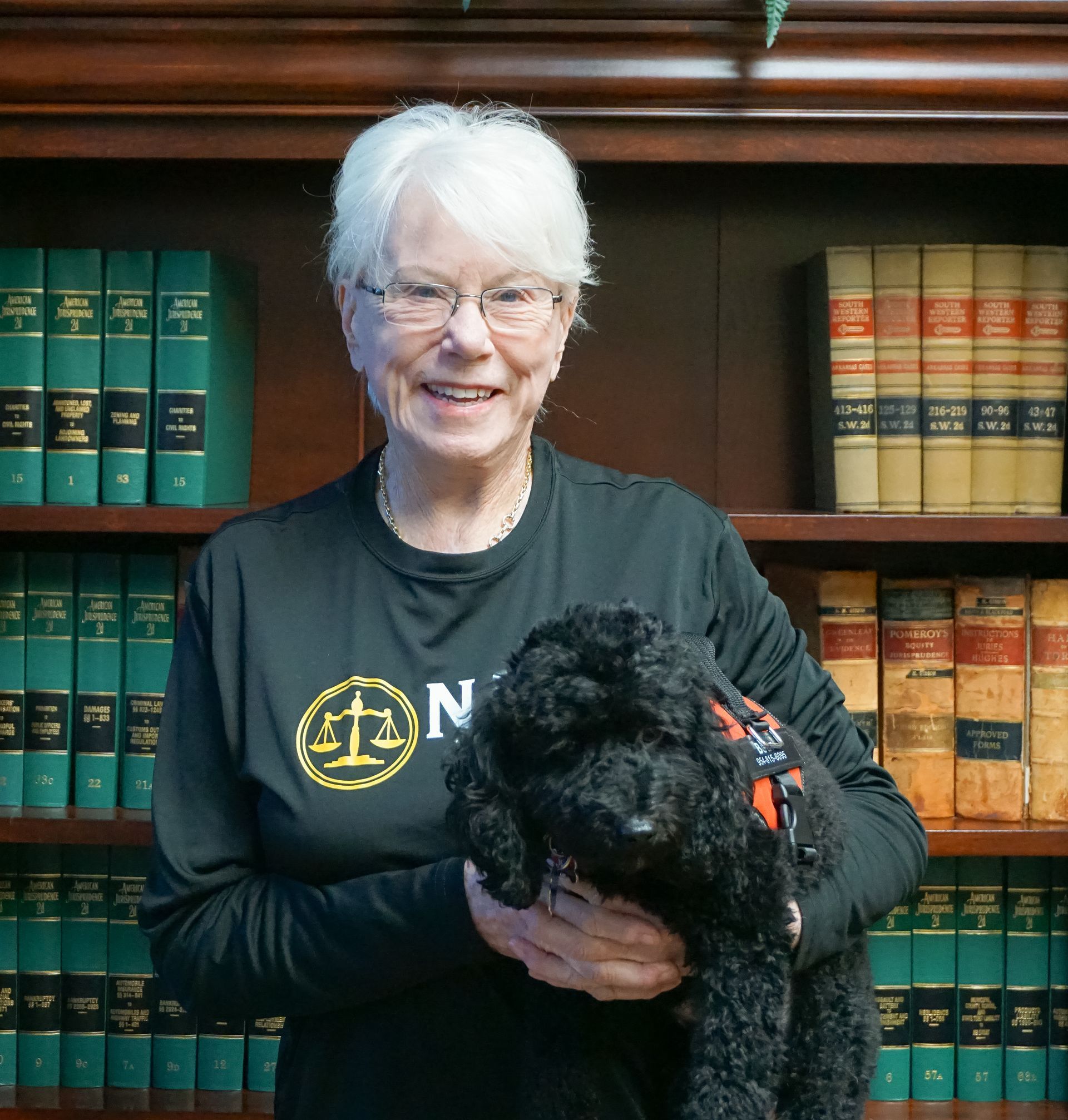 Woman with white hair holding a black dog in front of a bookshelf filled with books.