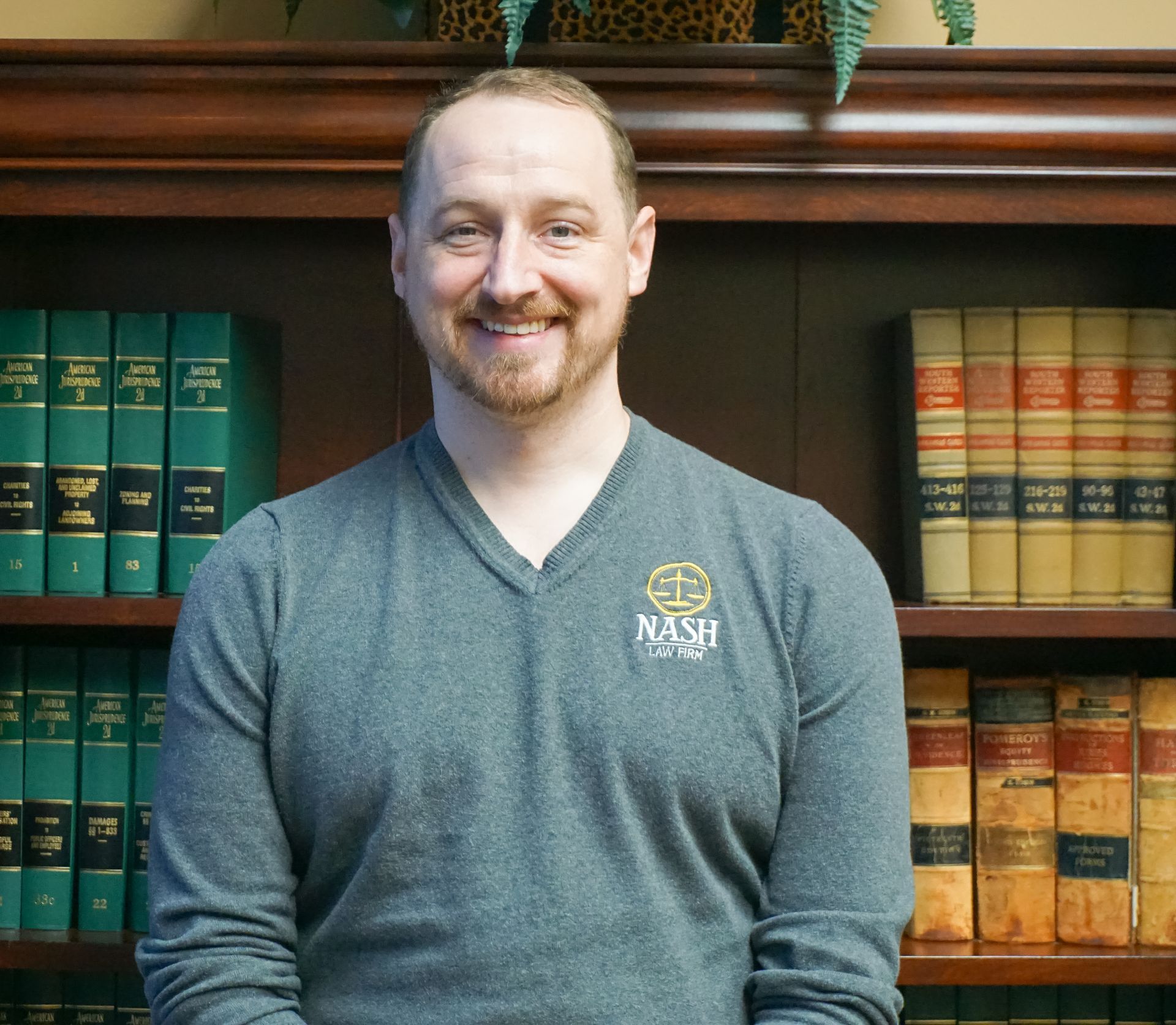 Man with short hair and beard wearing gray sweater in front of a bookshelf, smiling.