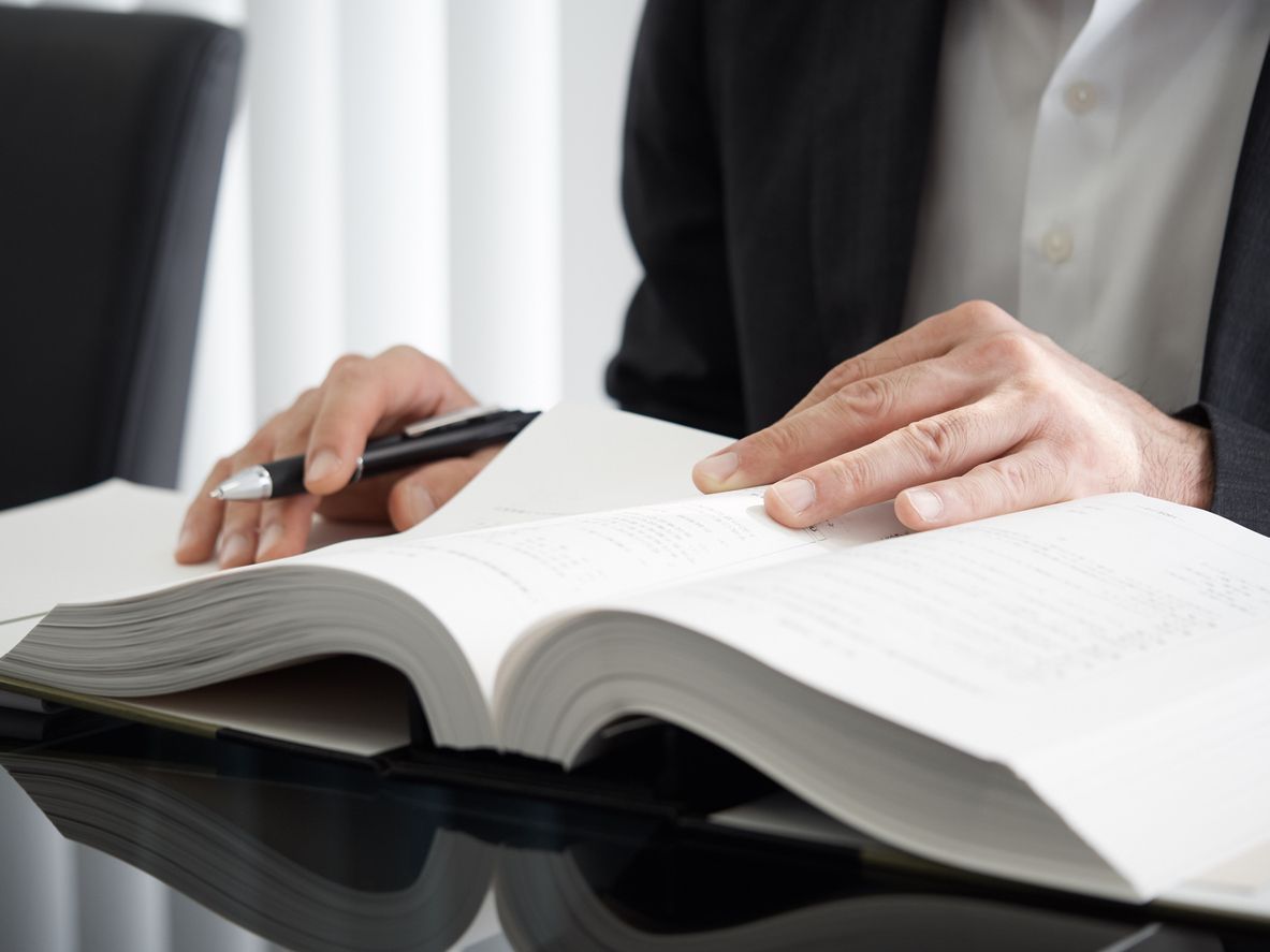 Person in a suit holding a pen and looking at an open book on a dark surface.