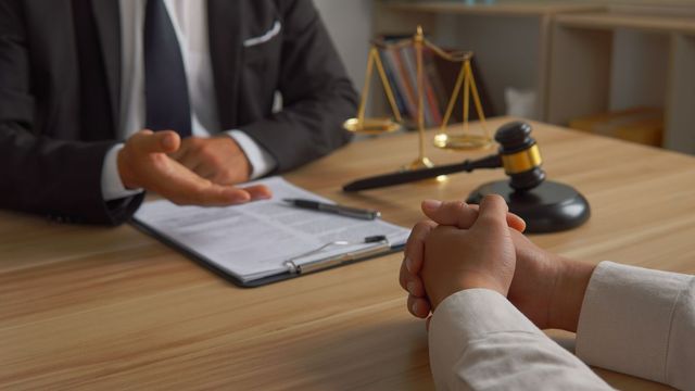 Lawyer gesturing to client; legal documents, gavel, and scales of justice on desk.