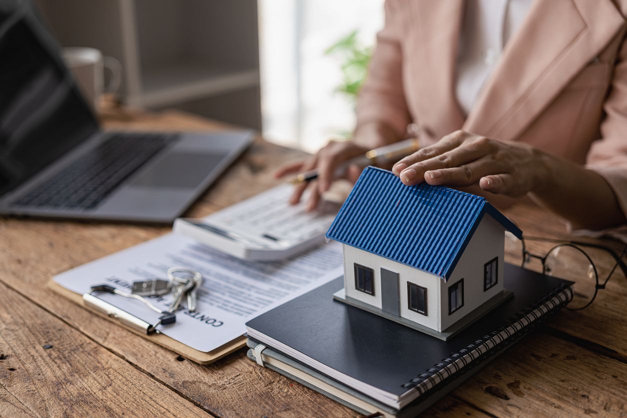 Person with hands on a model house on a desk with documents, a calculator, and a laptop.