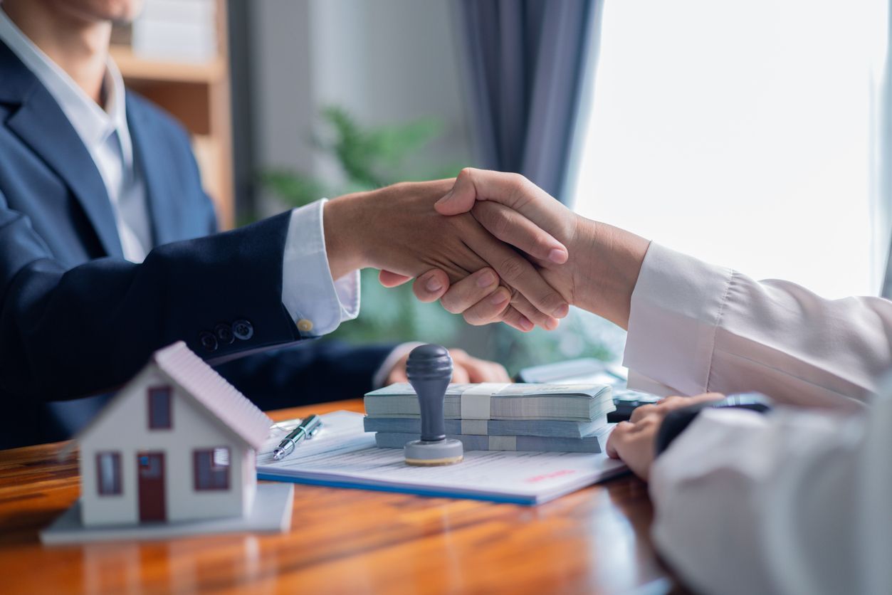 Two people shaking hands over paperwork and money, with a house model and stamp in the foreground.