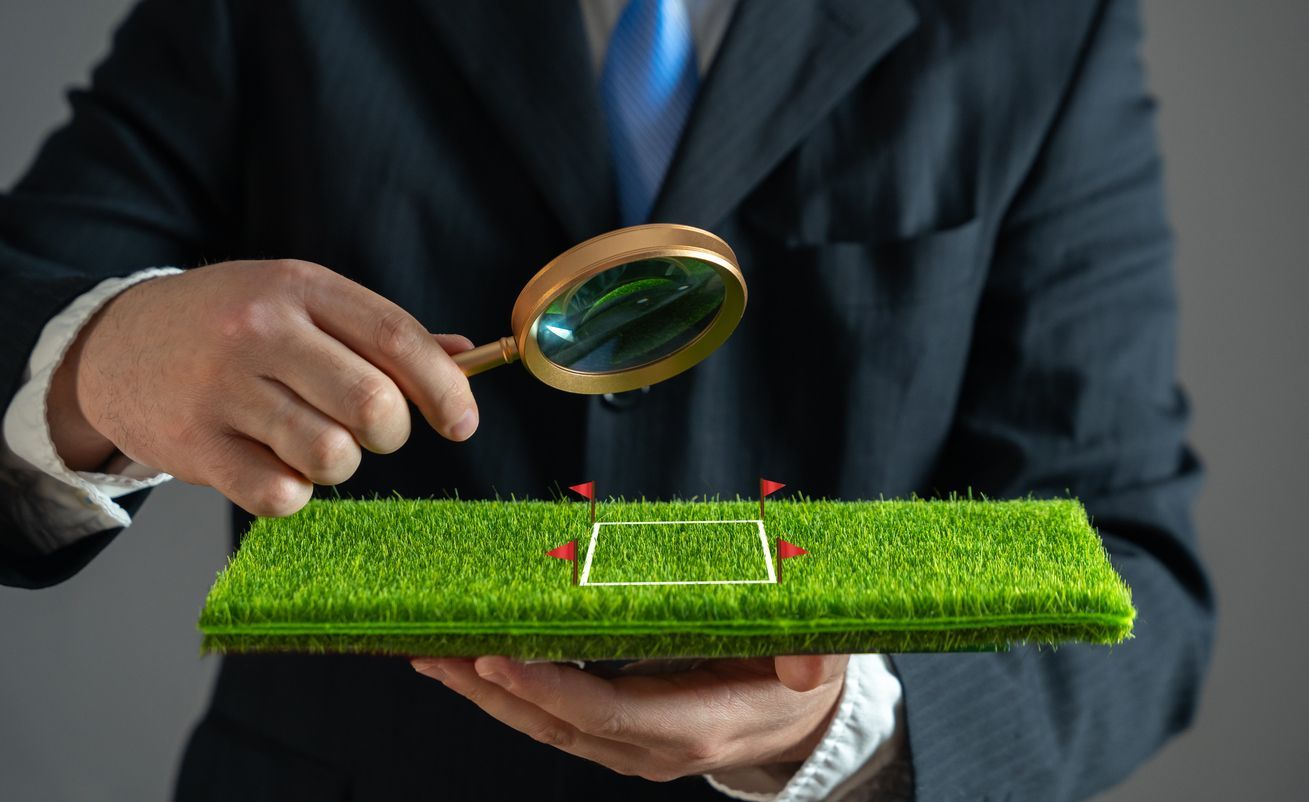 A person in a suit examining a miniature grass soccer field with a magnifying glass.