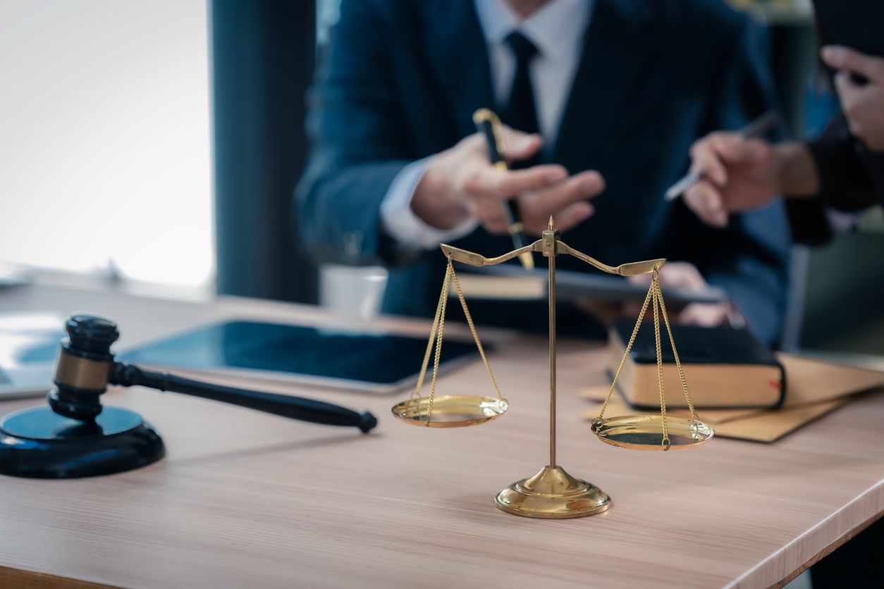 Scales of justice, gavel, and two people in suits at a desk, possibly lawyers, discussing documents.