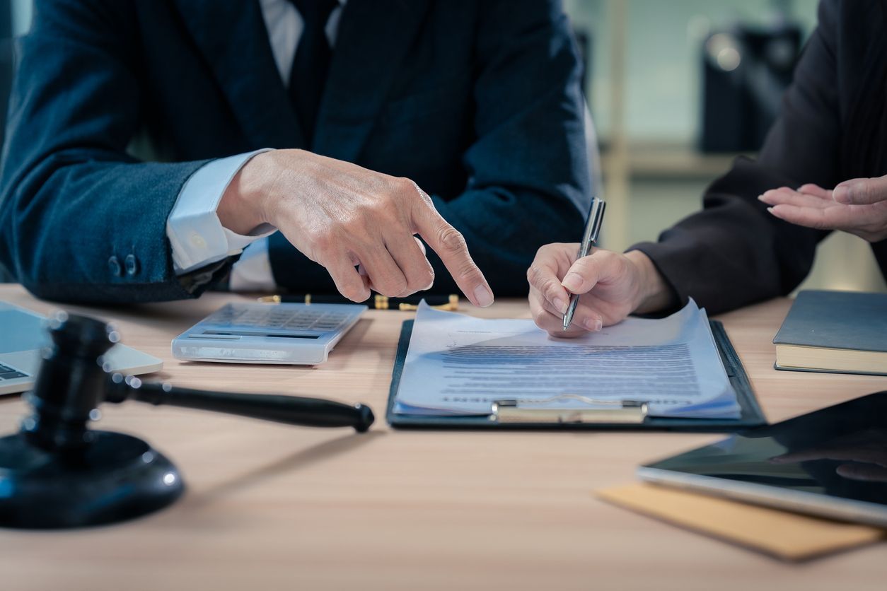 Two people in suits at a desk, one points, the other signs a document. Gavel and other legal items present.