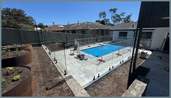 Backyard with pool enclosed by glass fence, surrounded by garden beds and patio. Blue sky.