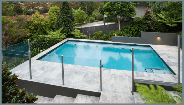 Rectangular swimming pool with blue water surrounded by gray stone and lush green landscaping.