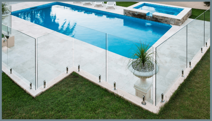 Glass pool fence surrounding a rectangular pool and jacuzzi on a lawn.