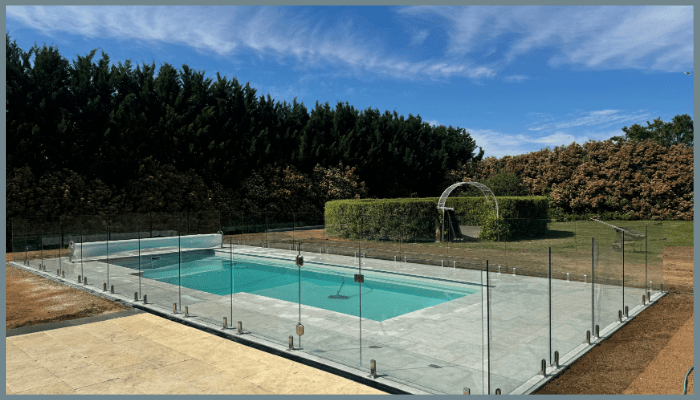 Pool with glass fence, surrounded by greenery and blue sky.