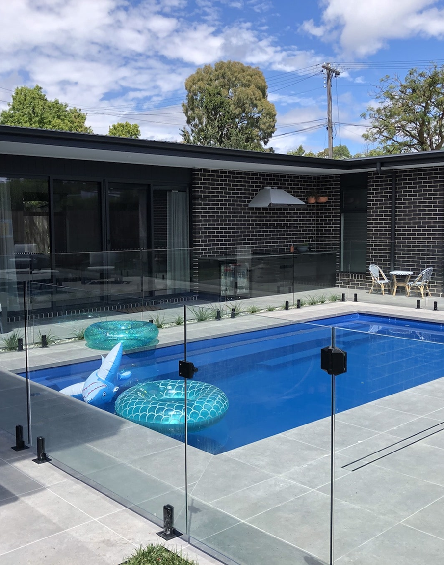 A large swimming pool with a glass fence and a house in the background.