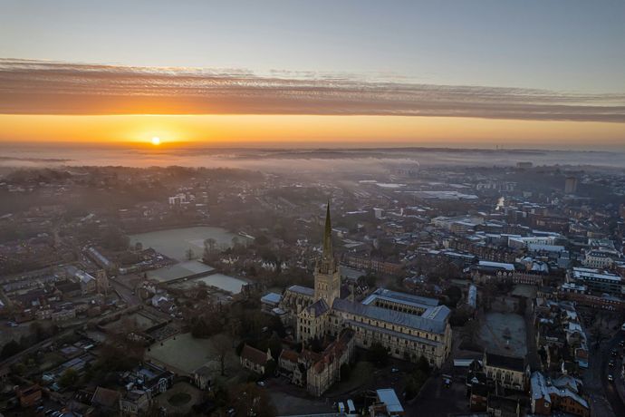 Aerial view of Norwich Cathedral at sunrise, the sun glowing orange over a hazy, fog-filled city.