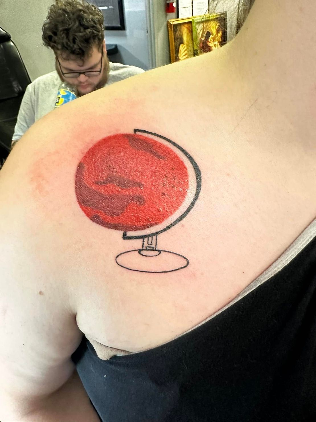 Red globe tattoo on a person's shoulder blade, with a black-outlined stand and red shading.