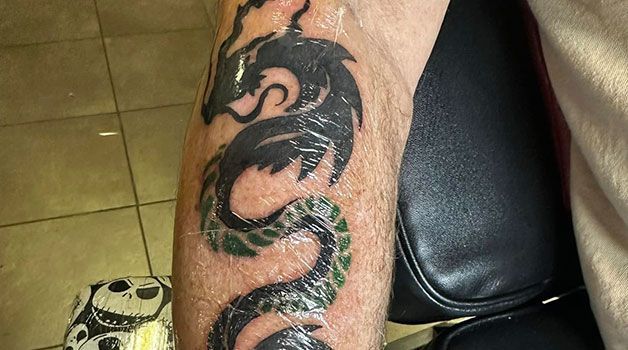 Black dragon tattoo on a person's forearm, with green accents on a bent arm near a black chair