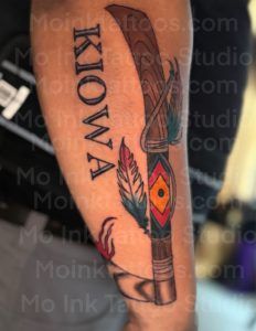 Tattoo of a canoe paddle with feather and geometric pattern, and the word “Kiowa” on skin
