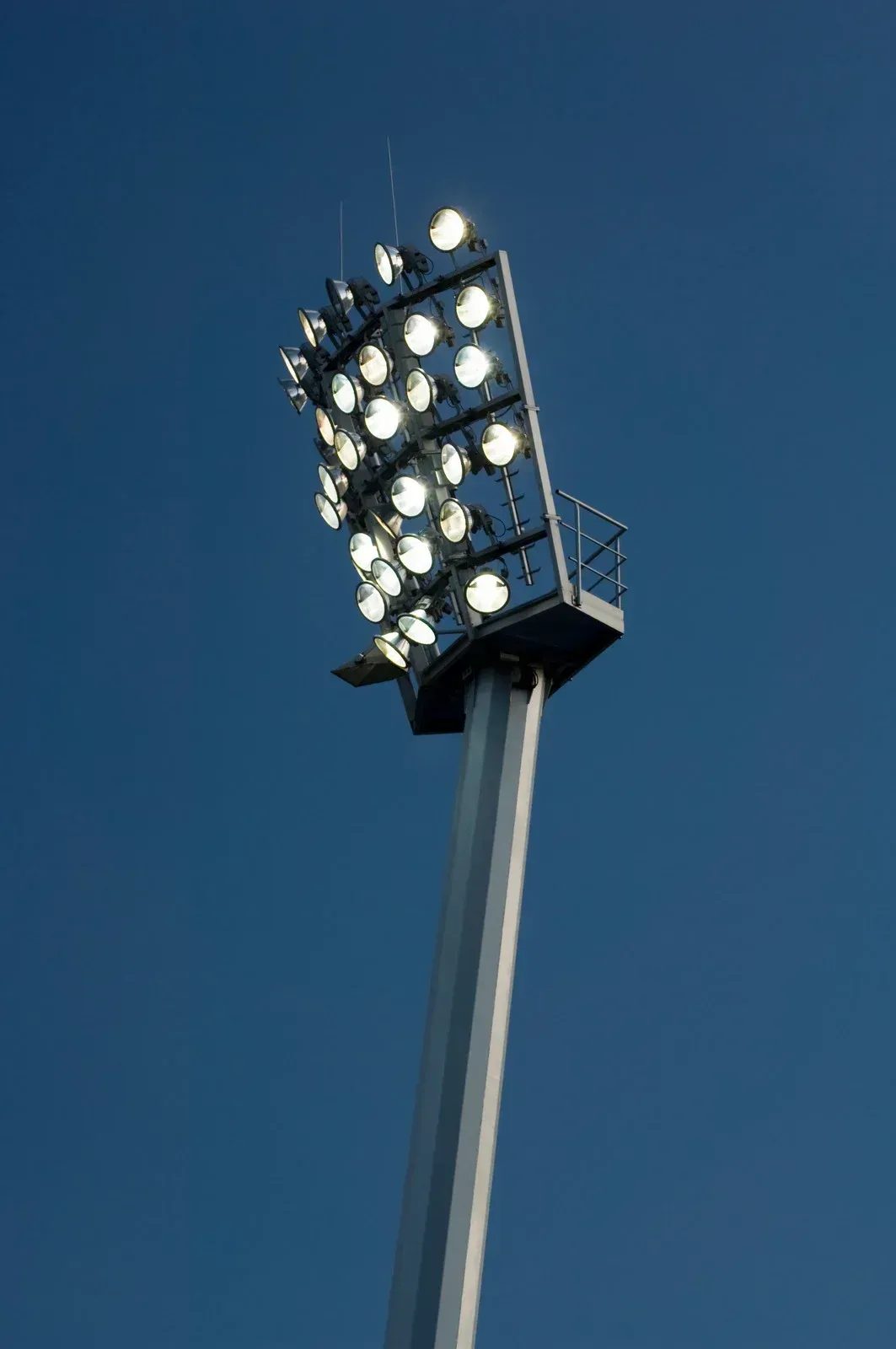 A stadium light is lit up against a blue sky.