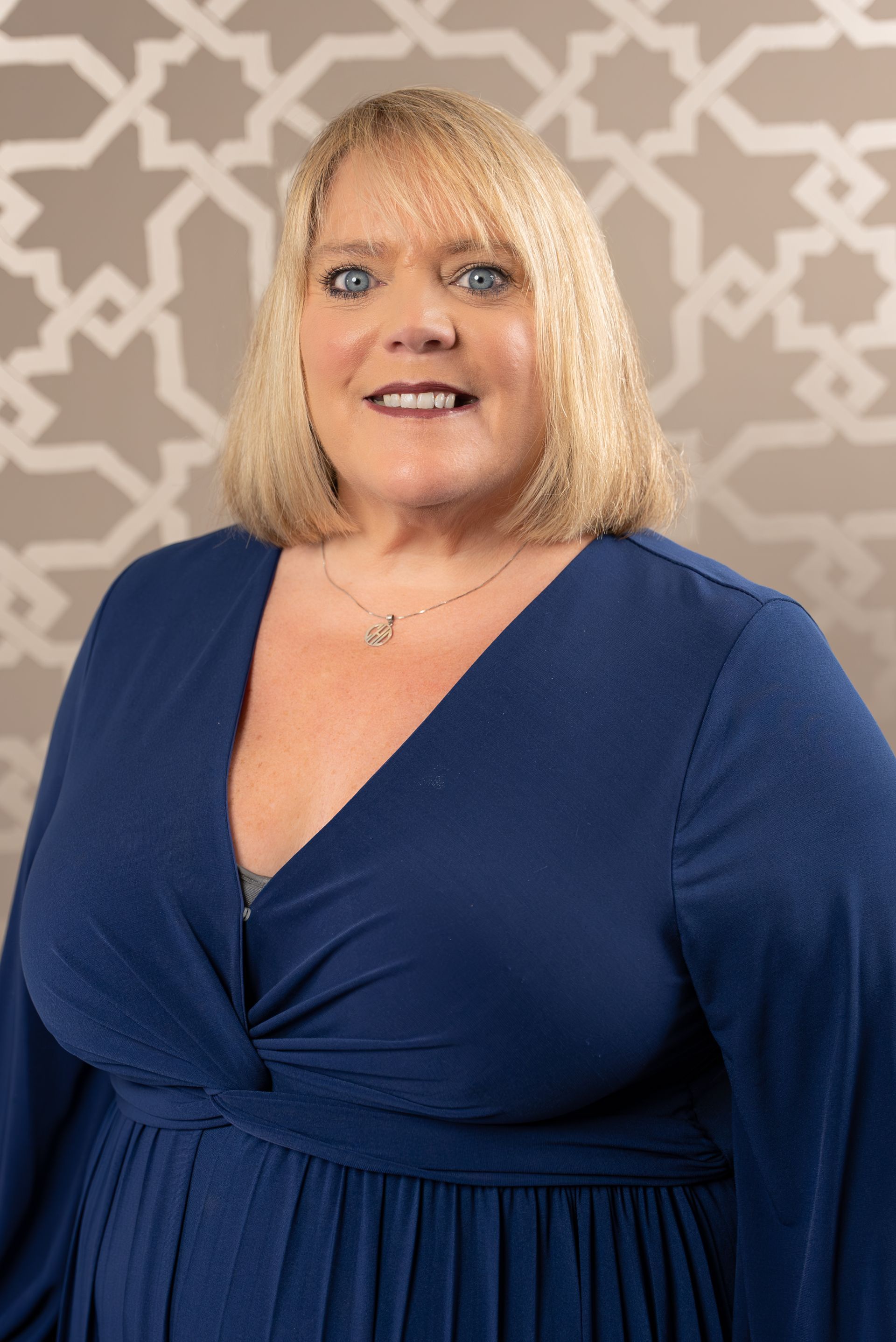 A woman in a blue dress is smiling in front of a patterned wall.