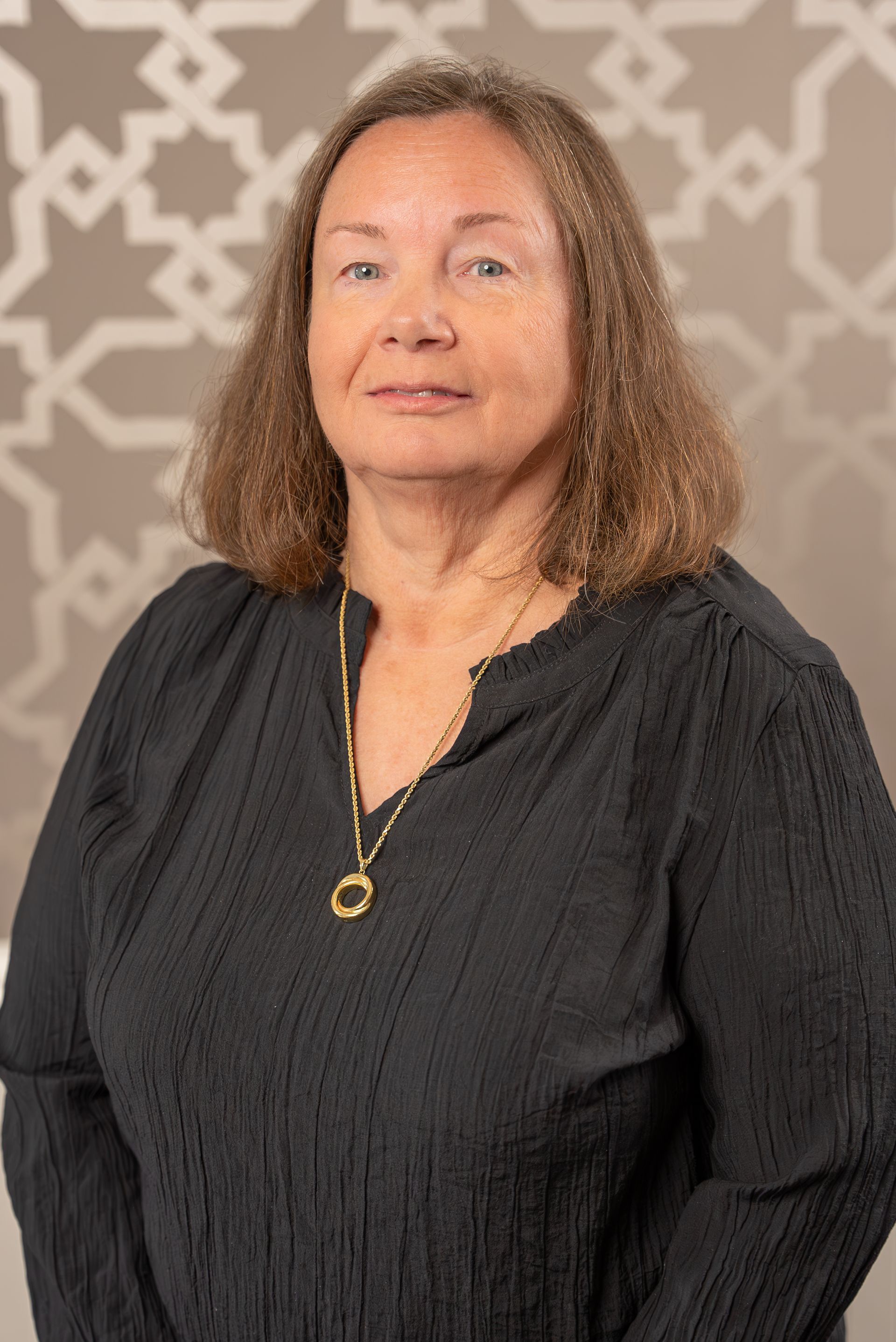 A woman wearing a black shirt and a gold necklace is standing in front of a patterned wall.