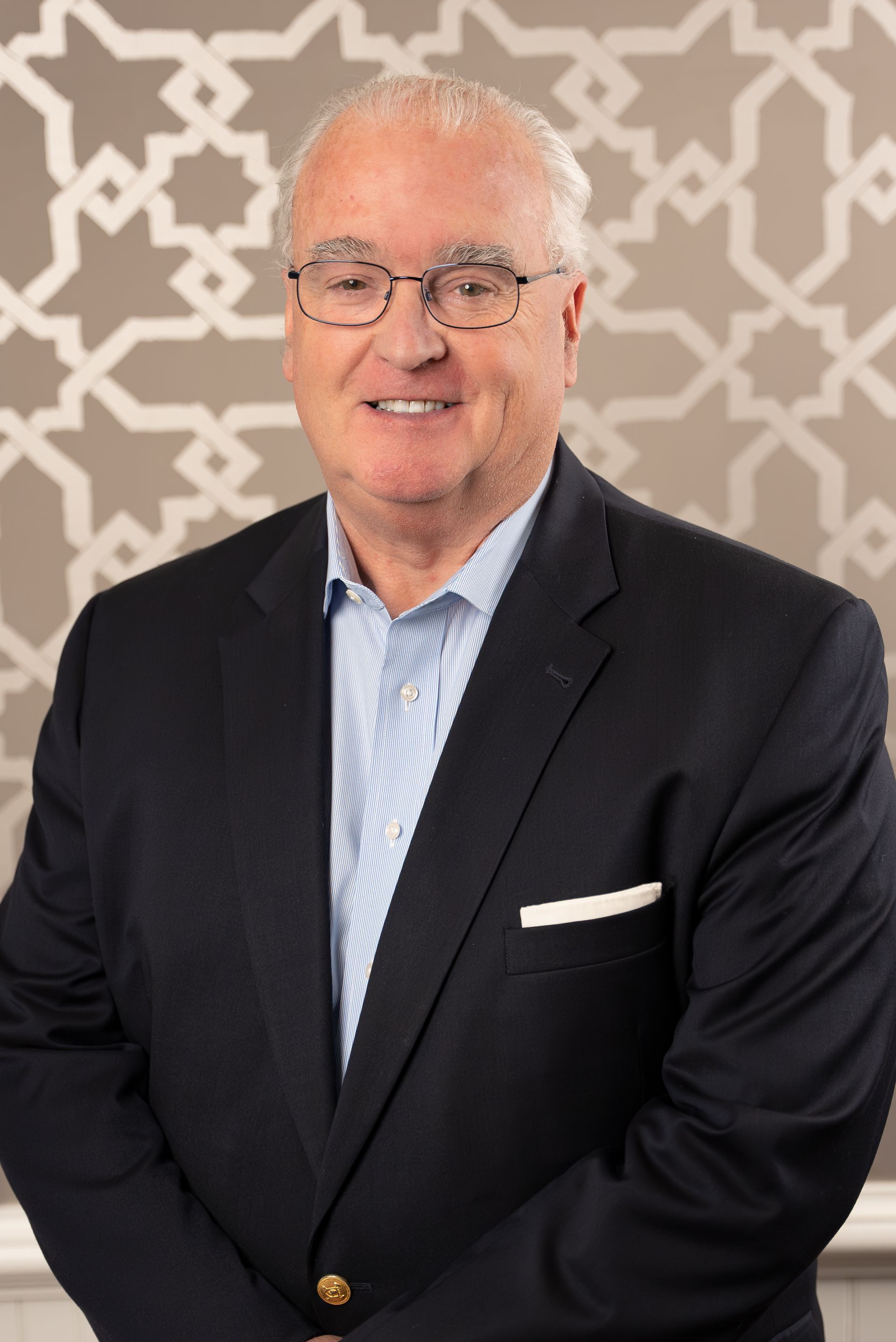 A man in a suit and glasses is standing in front of a wall.