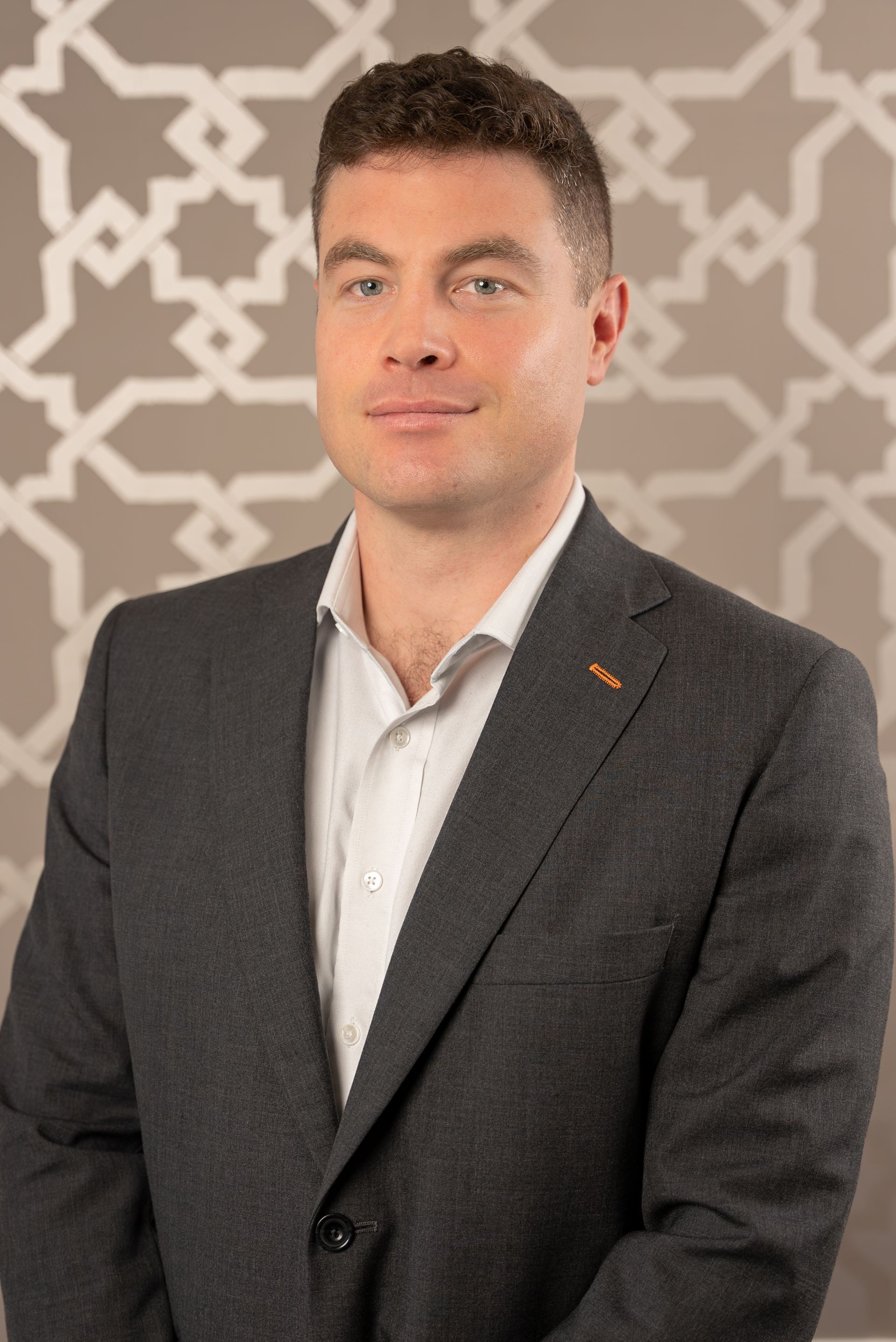 A man in a suit and white shirt is standing in front of a patterned wall.