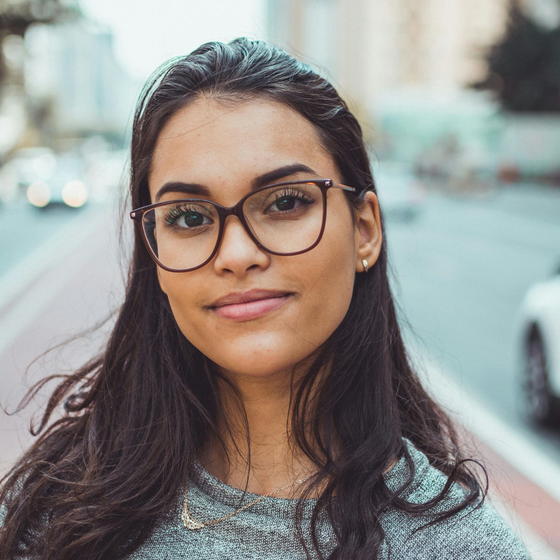 Woman with glasses smiling outdoors; blurred city background.