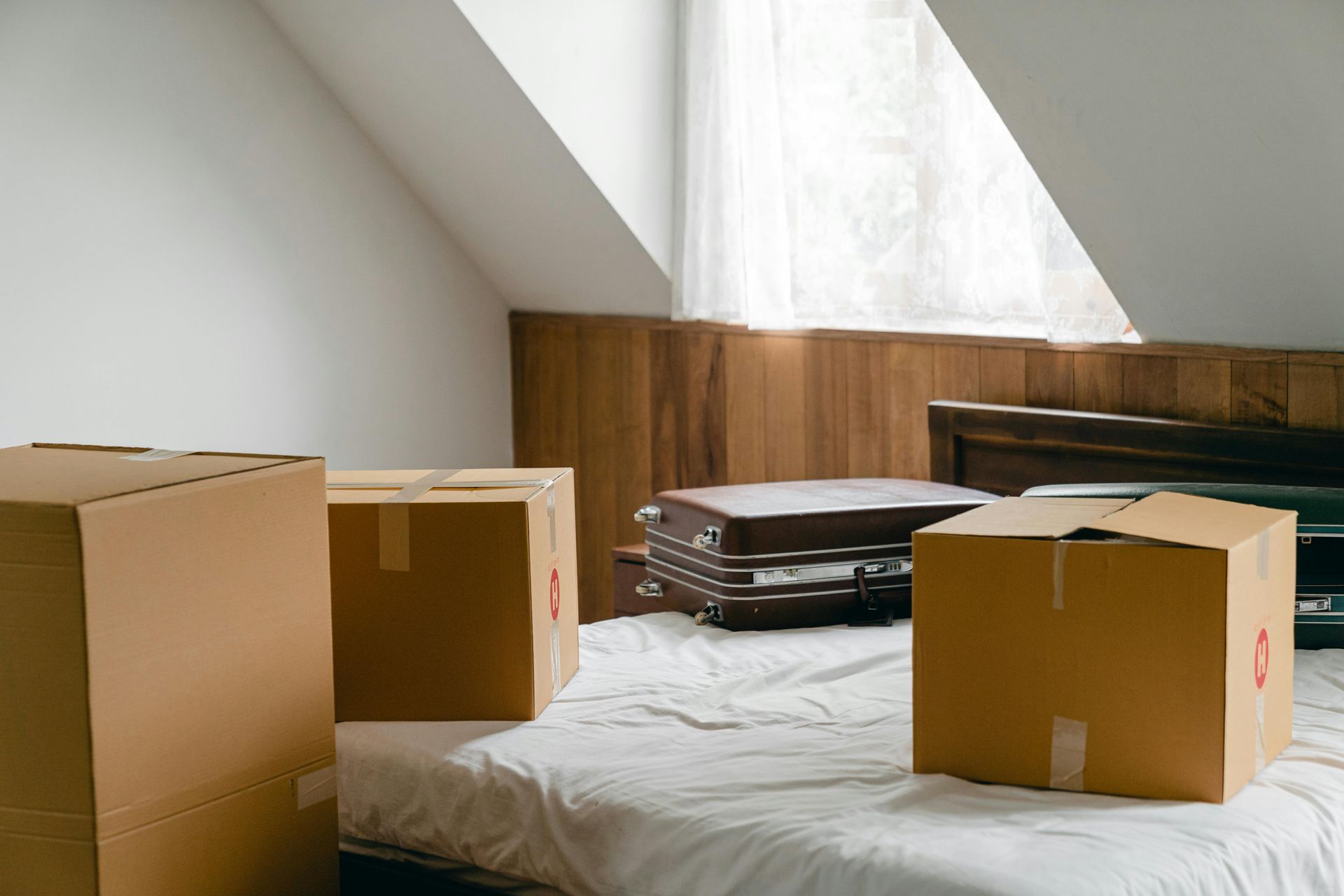 Three cardboard moving boxes and a vintage suitcase rest on an unmade bed in a room with slanted walls.