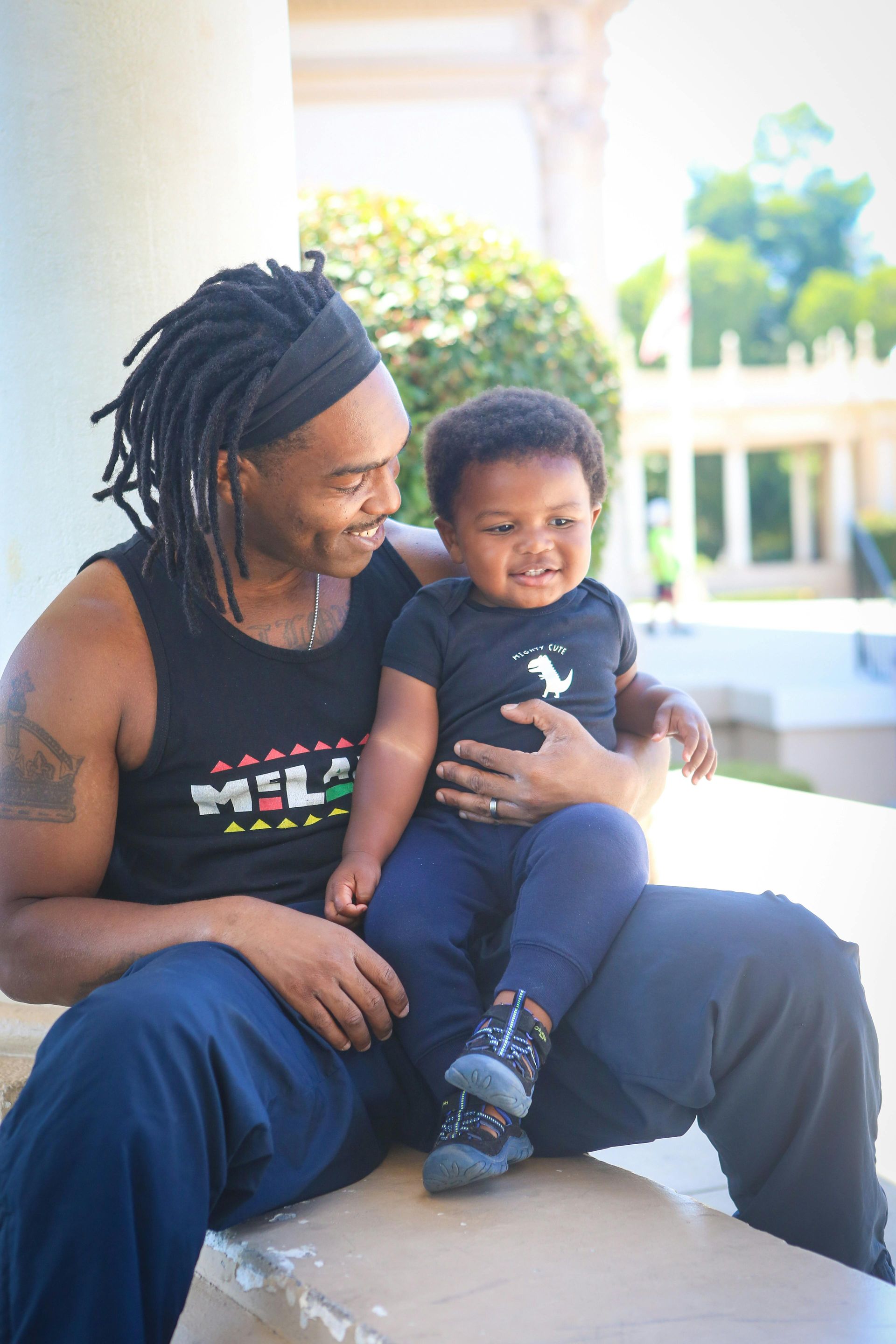 Man with dreadlocks holding a smiling toddler outdoors; both looking at each other, bright sunlight.