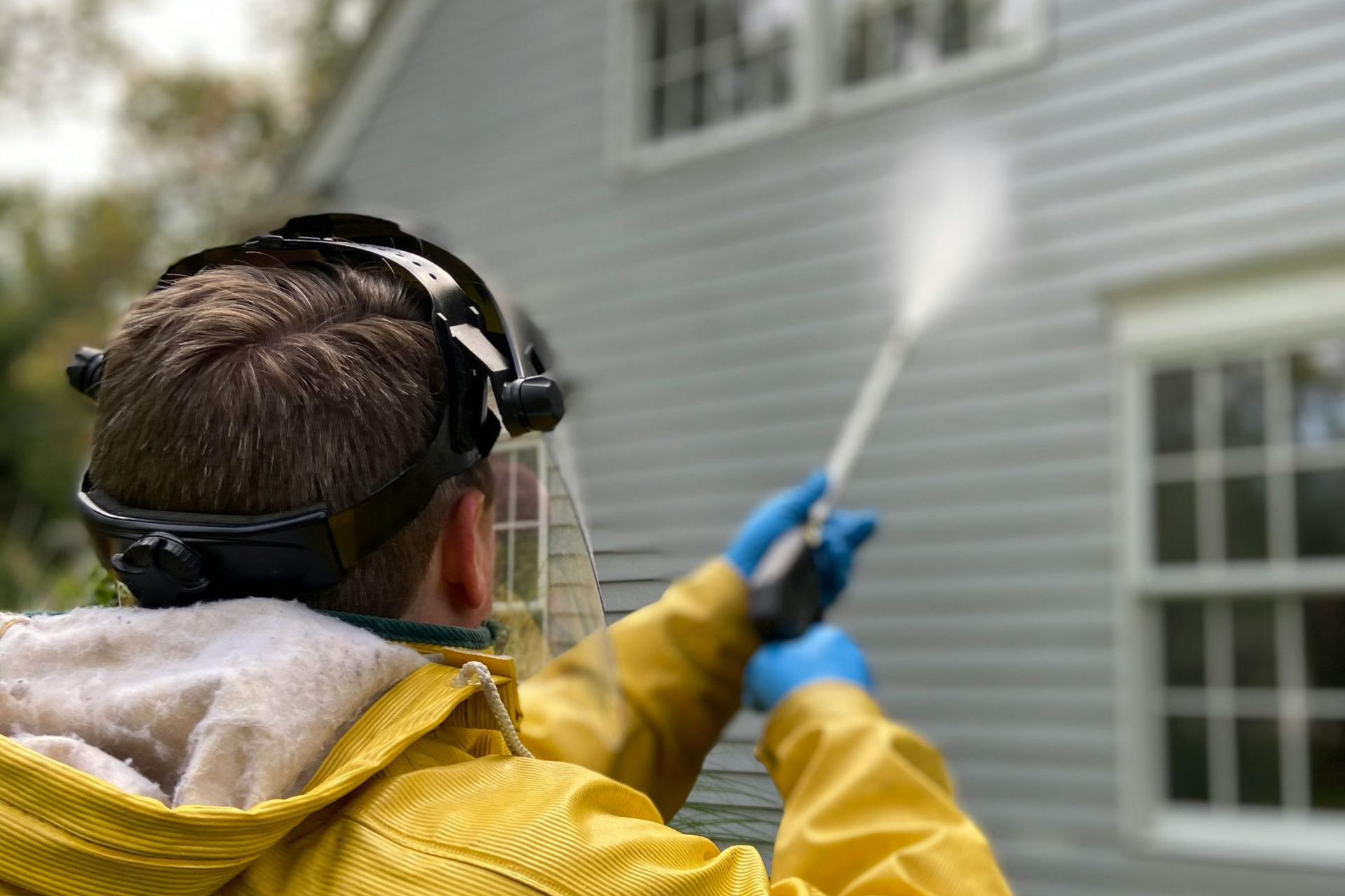 A person in a yellow jacket and protective face shield uses a pressure washer to clean the siding of a house.