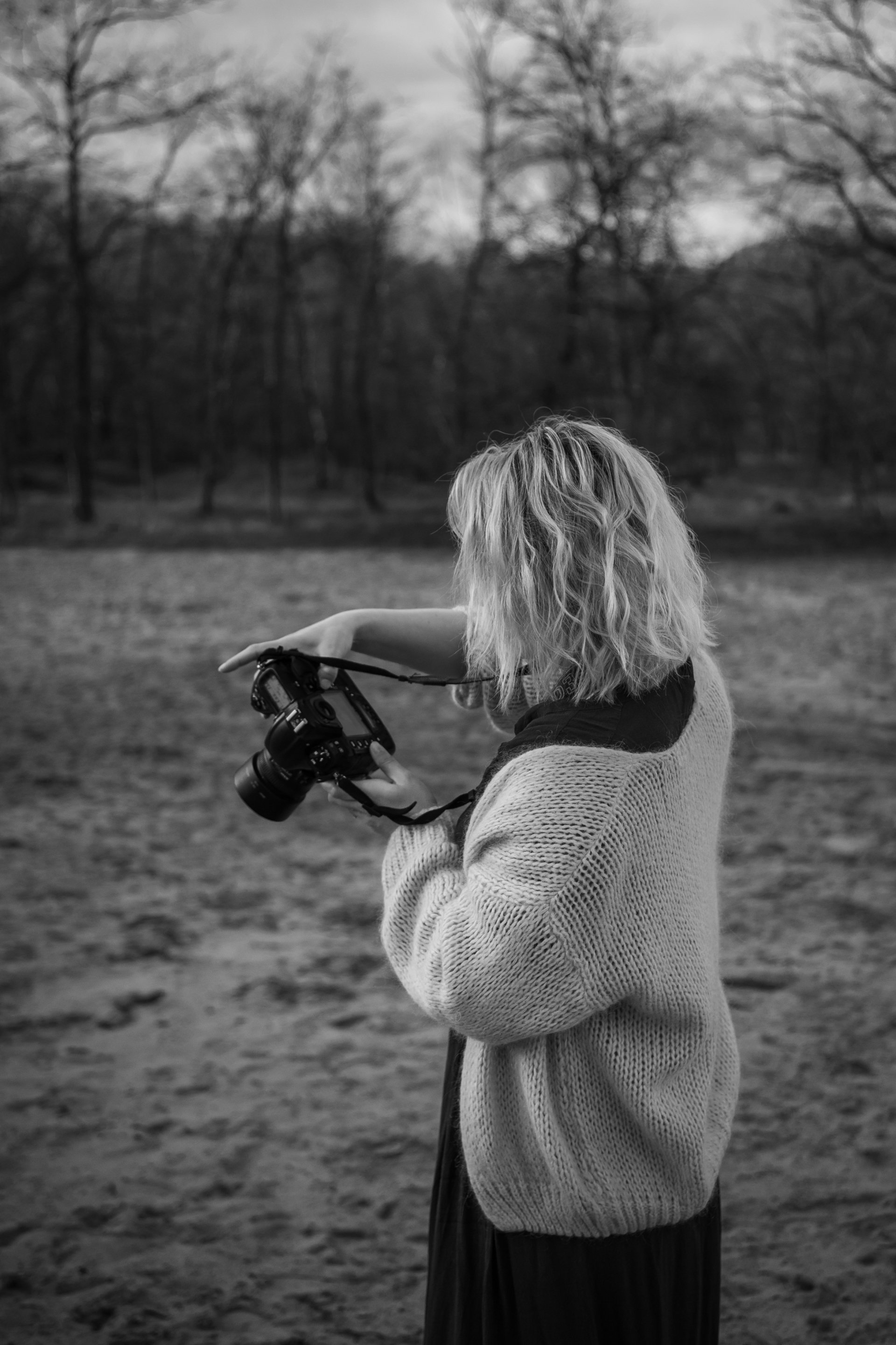 Vrouw met een camera in de hand kijkt buiten door de zoeker. Bomen en een veld op de achtergrond. Zwart-wit.