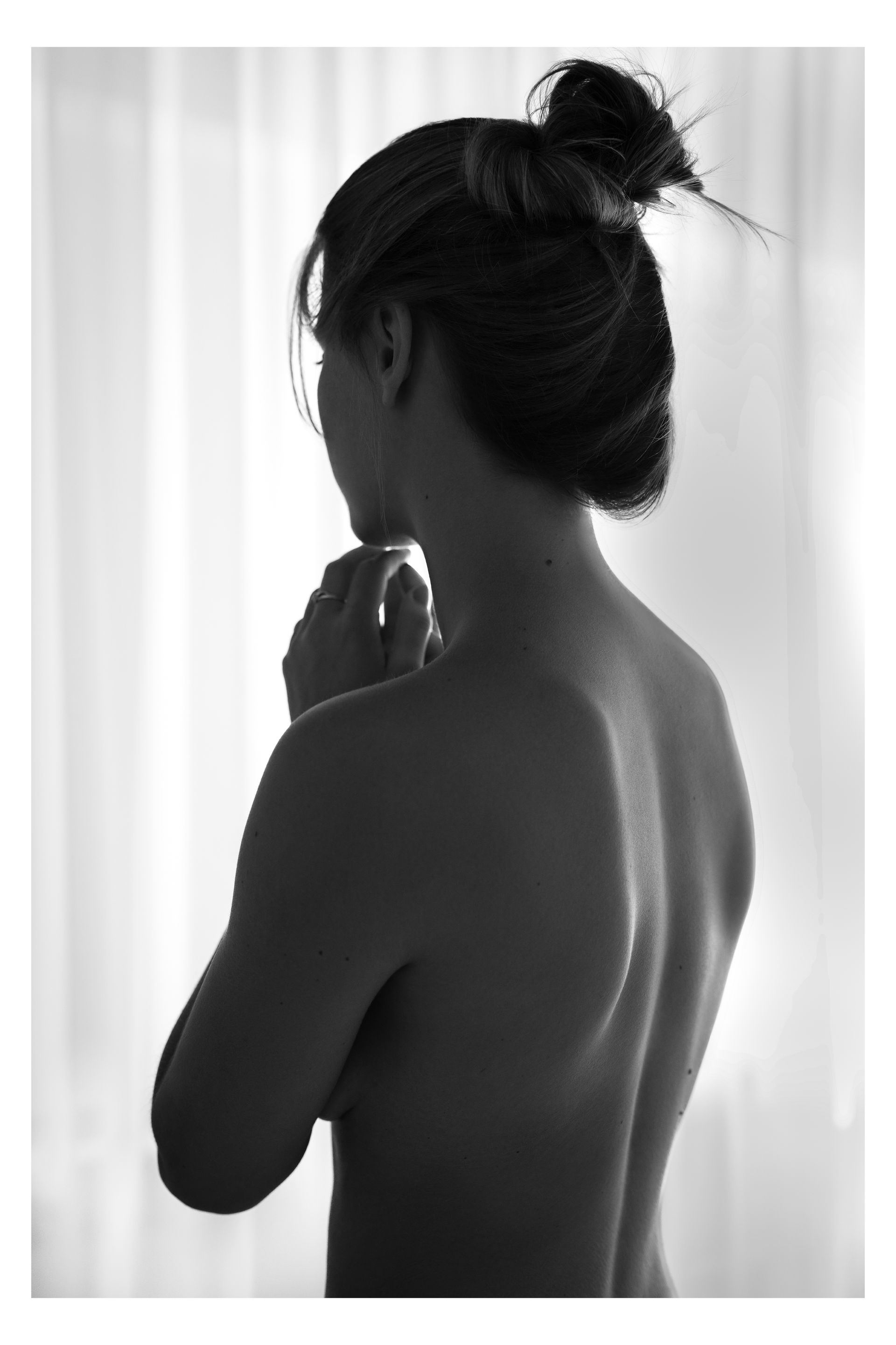 Silhouet van een persoon met opgestoken haar, met de rug naar de camera, handen ineengevouwen, voor een raam.