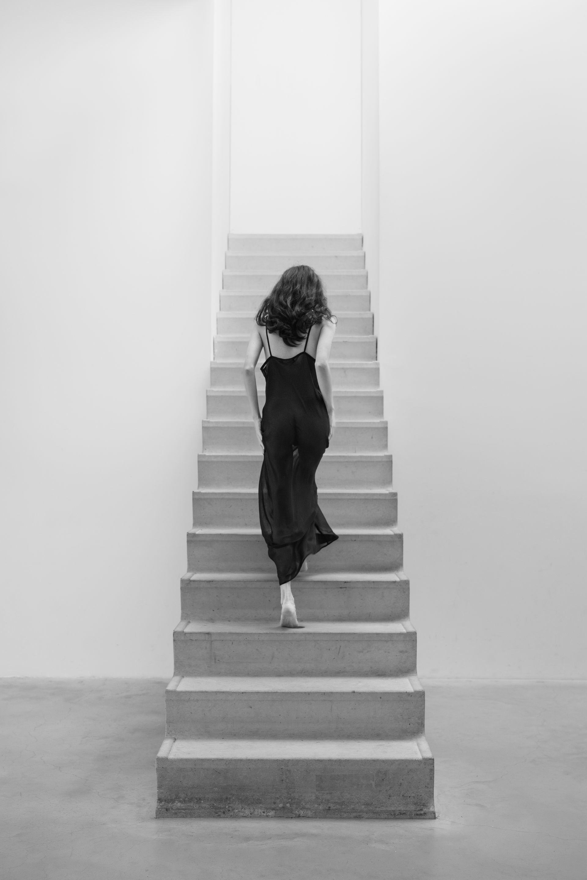 Vrouw in een zwart jumpsuit beklimt een betonnen trap in een witte kamer.
