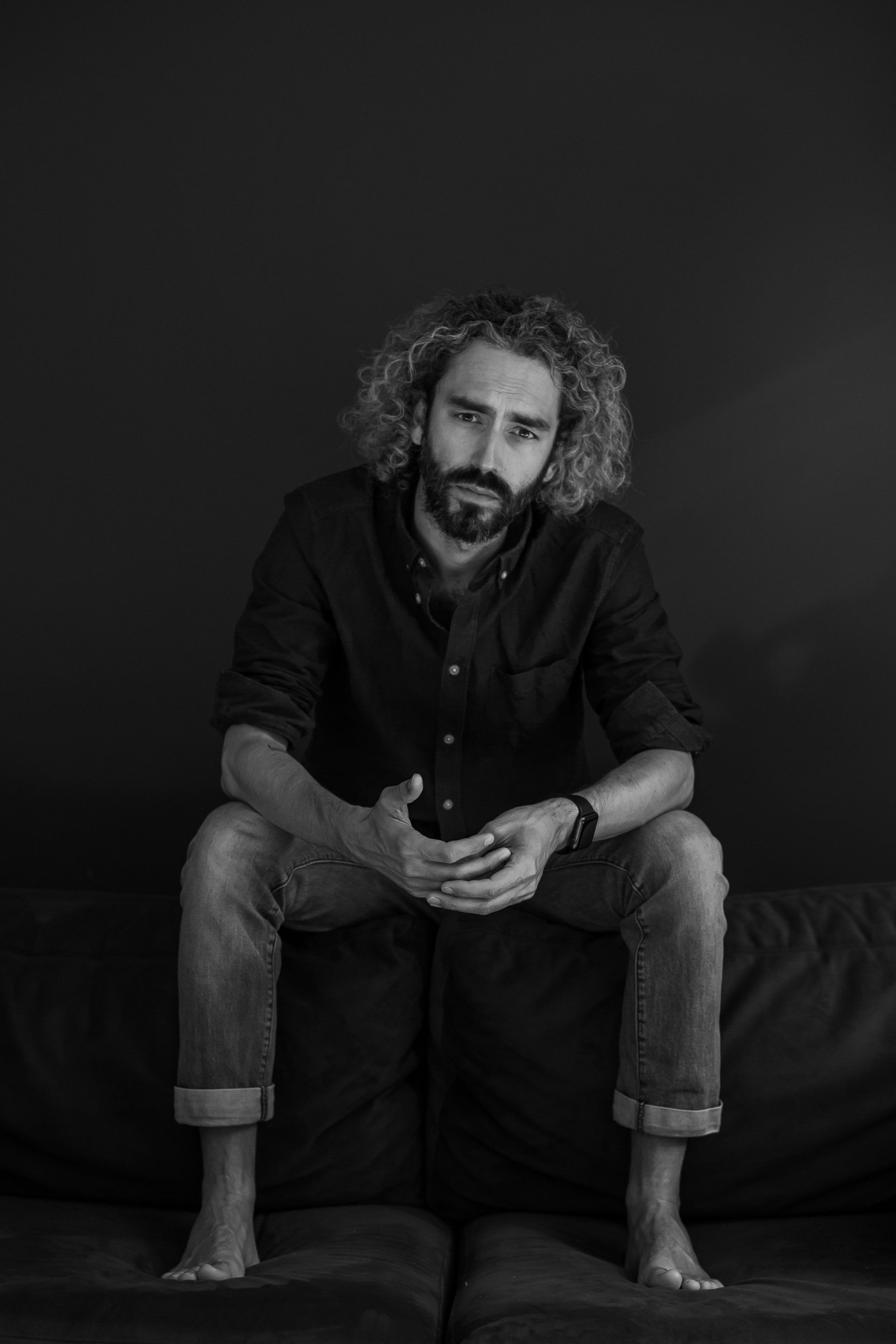 Een man met krullend haar, in een donker shirt en spijkerbroek, op blote voeten, zit op een bank, met de handen ineengevouwen en een serieuze uitdrukking op zijn gezicht.