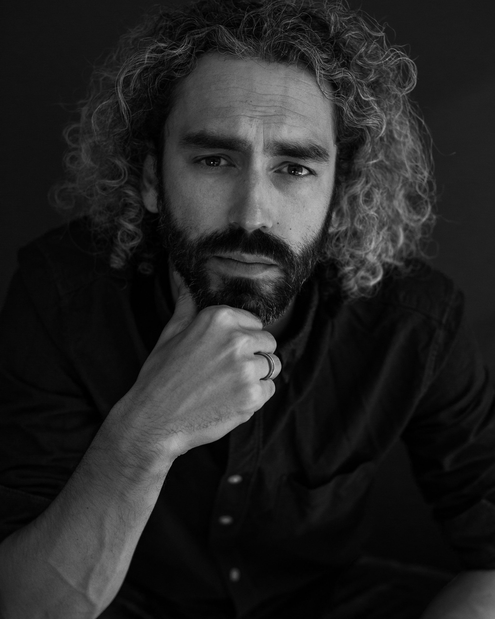 Man met krullend haar en een baard, die zijn kin op zijn hand laat rusten, kijkt in de camera.