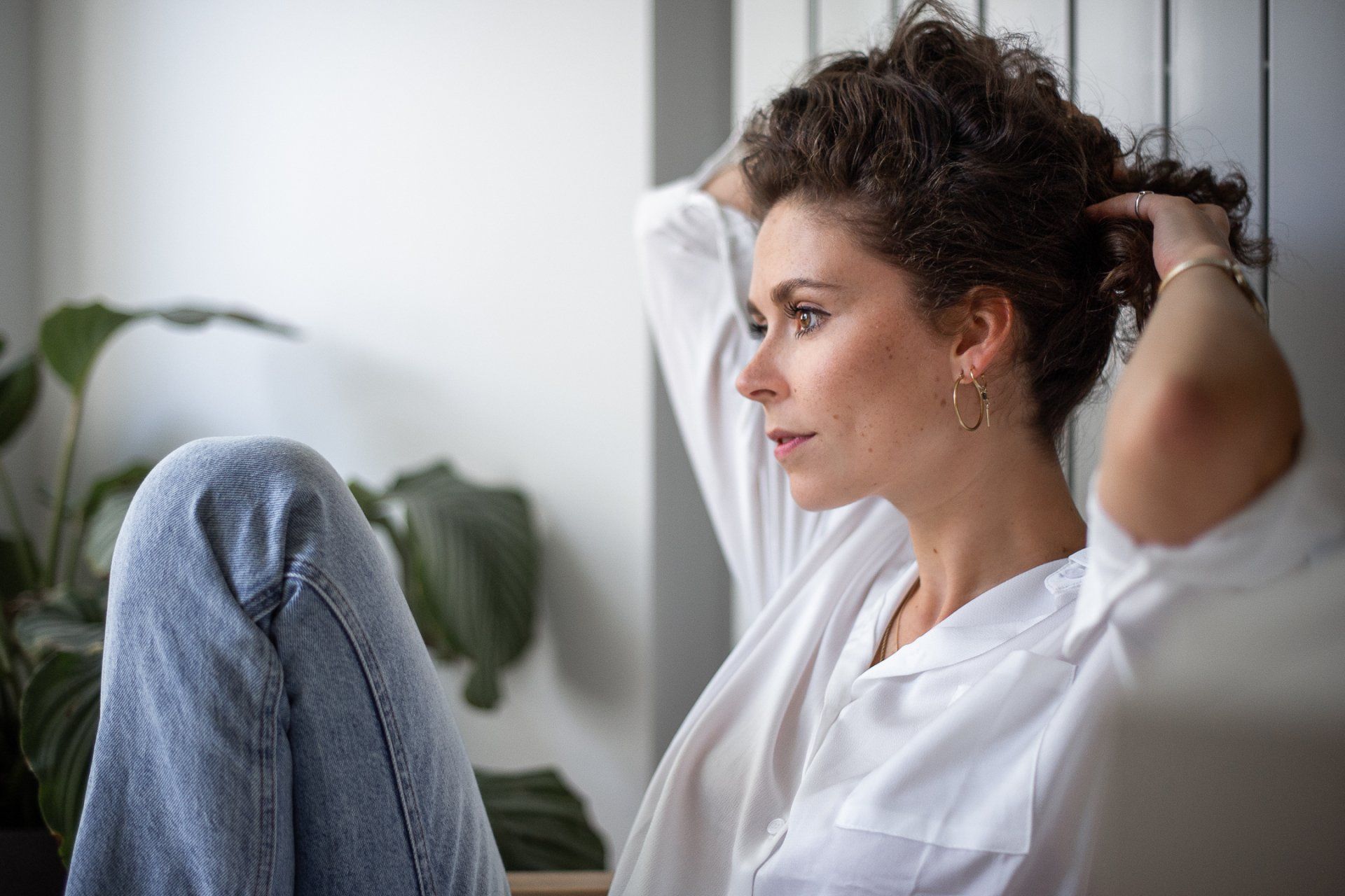 Vrouw met krullend haar, gekleed in een witte blouse en jeans, raakt haar haar aan bij een plant.