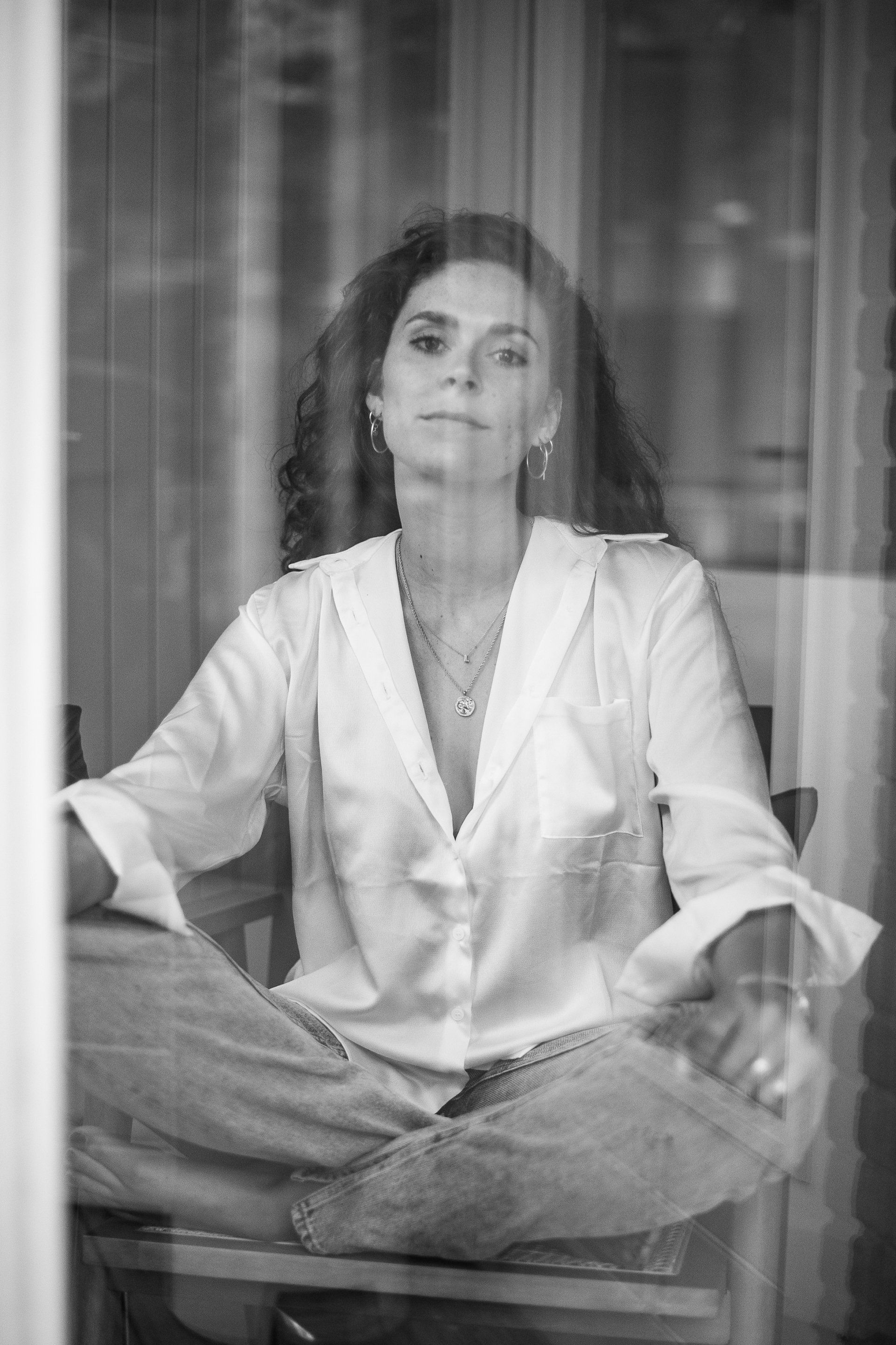 Een vrouw zit met gekruiste benen, gekleed in een witte blouse en spijkerbroek, en kijkt voor zich uit door een raam.