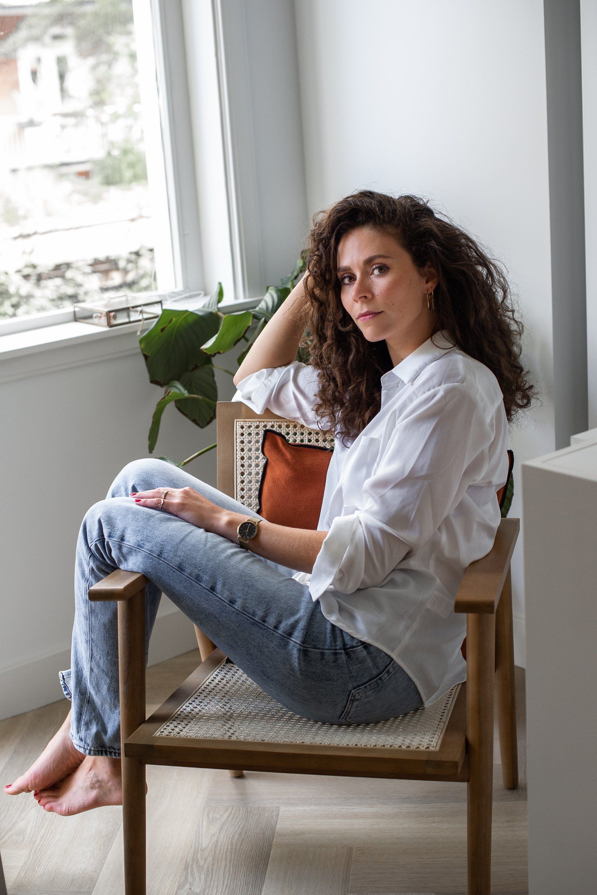 Vrouw met krullend haar, in een witte blouse en spijkerbroek, zit op een houten stoel bij een raam, met een plant.