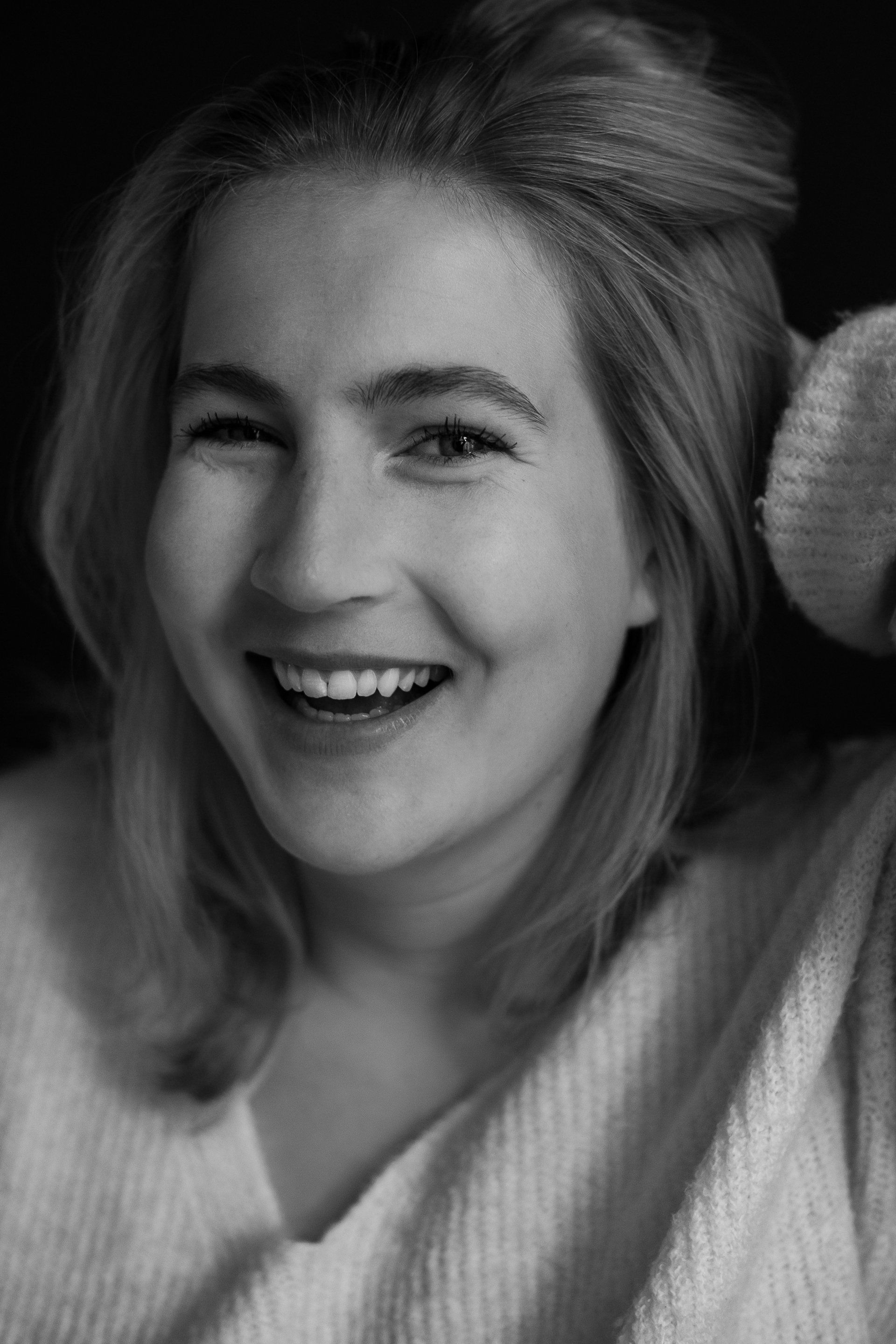 Vrouw met een brede glimlach, licht haar, draagt ​​een trui en houdt haar haar vast, tegen een donkere achtergrond.