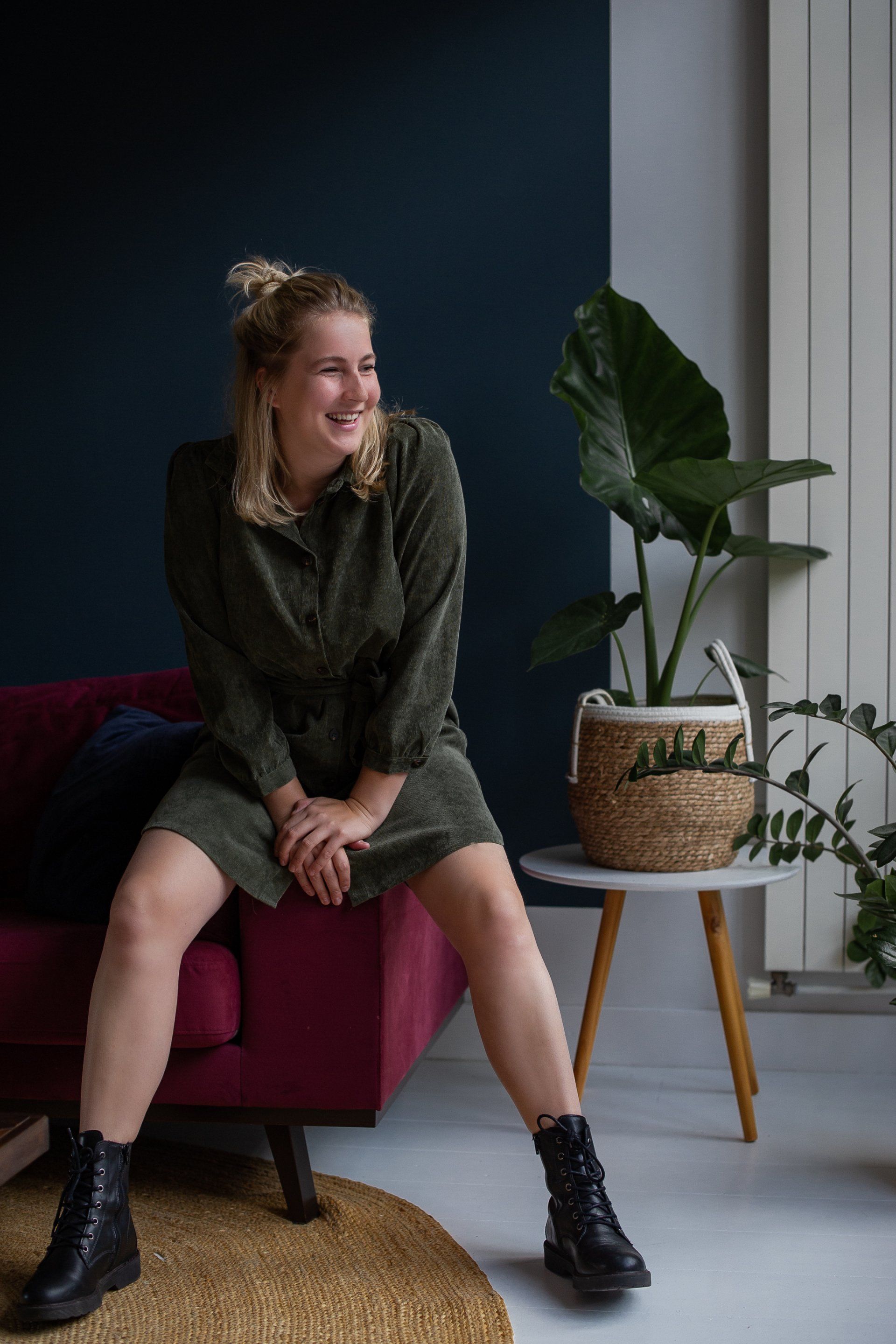 Een vrouw zit glimlachend op een bordeauxrode bank. Ze draagt ​​een groene jurk en zwarte laarzen, met een plant rechts van haar.