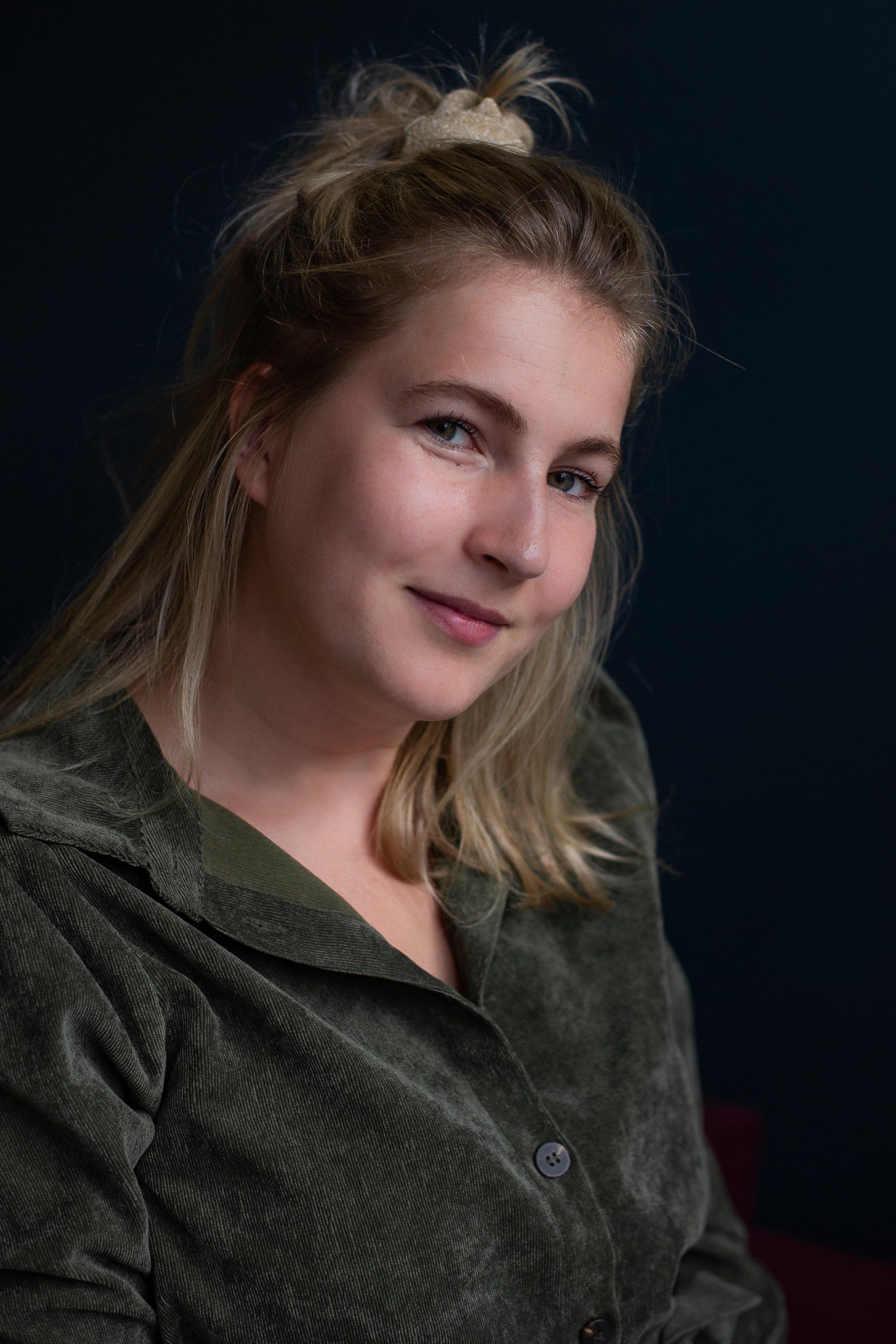 Een vrouw met blond haar in een knot, gekleed in een groene corduroy blouse, glimlacht lichtjes tegen een donkerblauwe achtergrond.